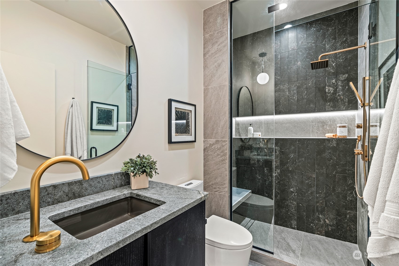 Elegant bathroom featuring a modern glass-enclosed shower with sleek black tiles, gold fixtures, and integrated lighting. Includes a stylish countertop, vessel sink, and a chic round mirror for a luxurious touch.