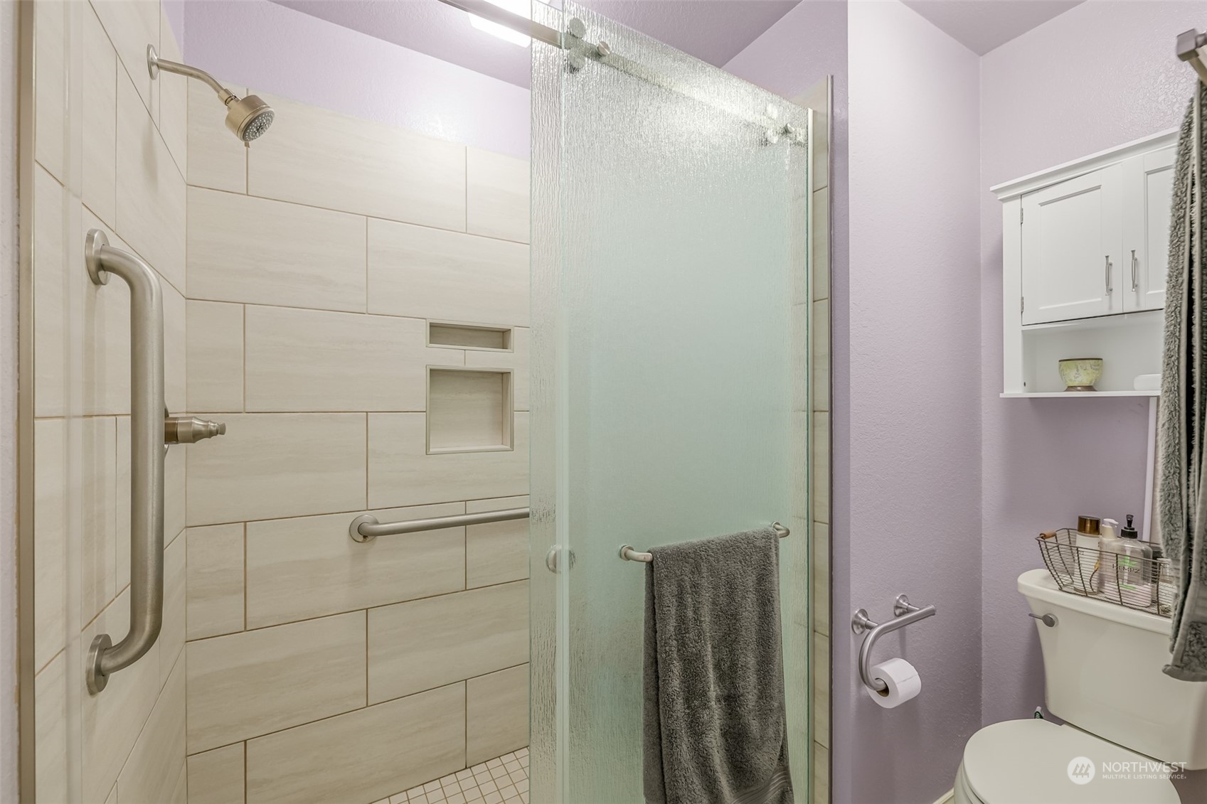 This beautifully tiled walk-in shower offers both style and accessibility, featuring a sleek glass door and safety grab bars. The combination of contemporary fixtures and thoughtful design ensures comfort and convenience.