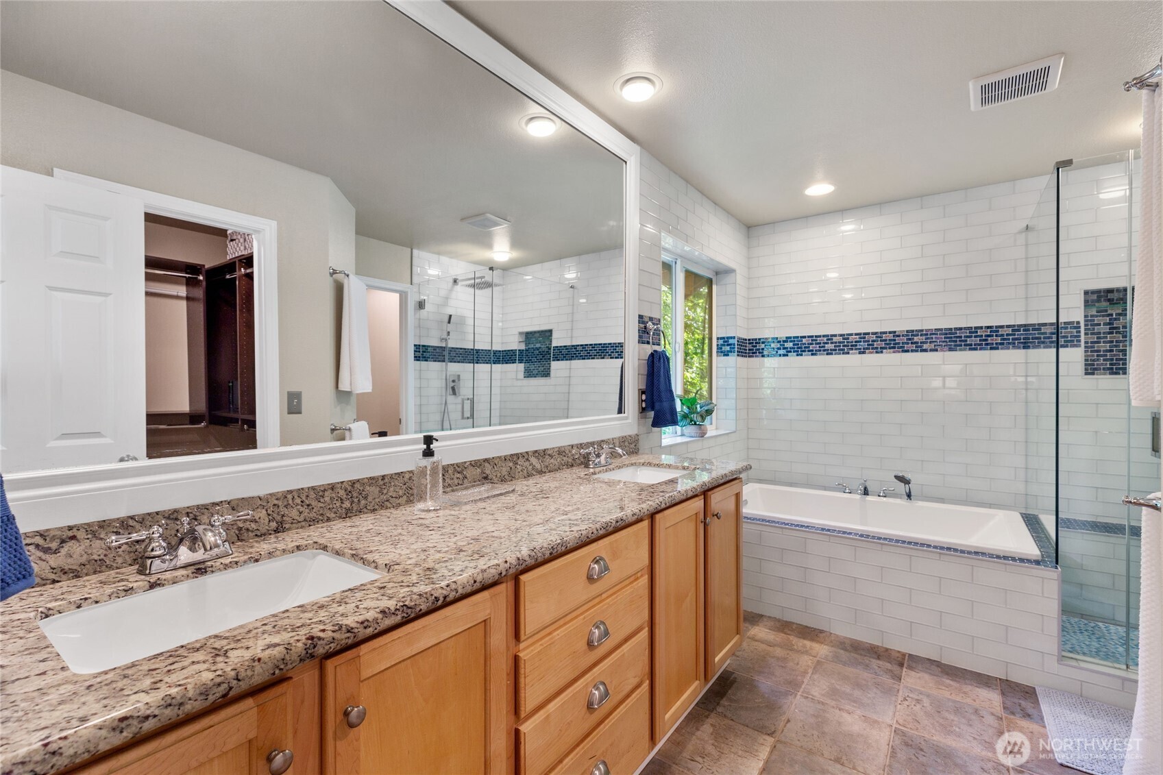 Primary bathroom with a double vanity, granite countertops and a mix of white subway and blue accent tiles. Features a soaking tub by the window, glass-enclosed shower, and natural light throughout.
