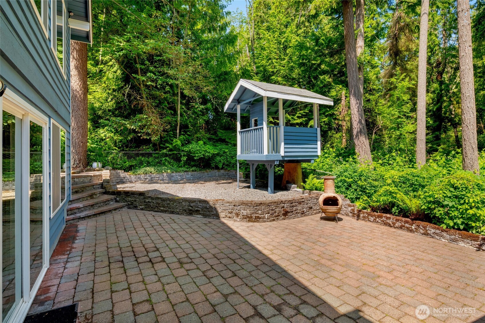 Expansive paver patio bordered by lush greenery offers a serene space to gather, dine, or play. The charming covered structure is perfect for a playhouse, potting shed, or creative outdoor nook.