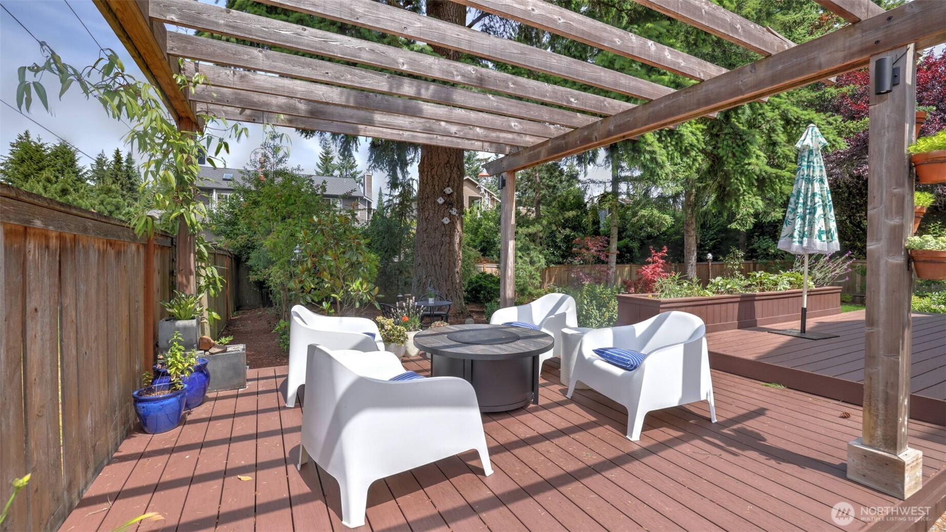 This spacious wooden deck showcases a charming two-tiered pergola, offering room for cozy seating and a fire pit. Surrounded by hanging greenery and perfect for an array of potted plants, it’s a serene outdoor retreat ready to be personalized