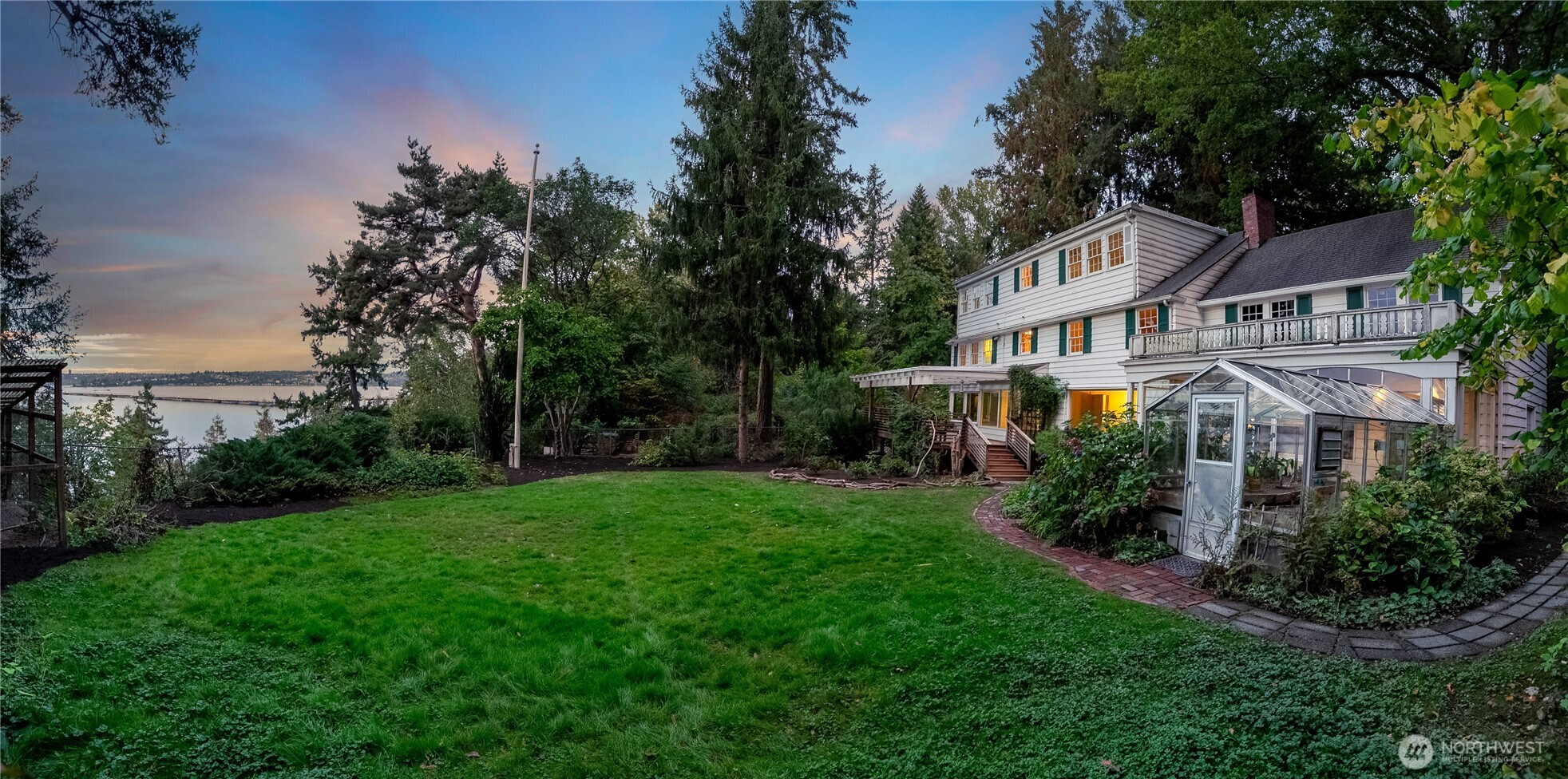 Framed by tall evergreens and the glimmer of Lake Washington beyond, this generous lawn offers a rare expanse of open space along the shoreline—a natural amphitheater for gatherings, games, or quiet contemplation. The historic home anchors the scene, its classic lines softened by trellises and trailing vines, while an open pergola and glass conservatory invite indoor-outdoor living. As dusk settles over the water, the entire setting glows with possibility—a place to linger, celebrate, and imagine what comes next.