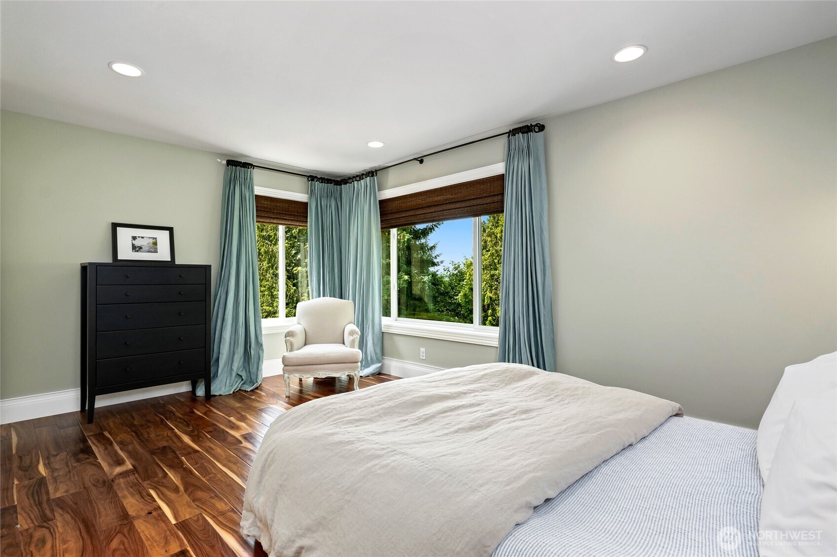Sunlit second lower level bedroom retreat with rich hardwood floors, peaceful green-toned walls, and corner windows offering treetop views and tranquil natural light.