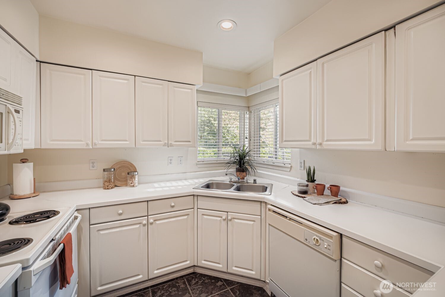 This bright and functional kitchen offers a clean, timeless design with crisp white cabinetry and matching appliances. A corner window above the double sink fills the space with natural light, creating a cheerful atmosphere for meal prep or morning coffee. Ample counter space and storage make this kitchen both practical and inviting, while subtle decorative touches add warmth and charm. Perfectly positioned for everyday convenience, this kitchen blends classic style with effortless functionality.