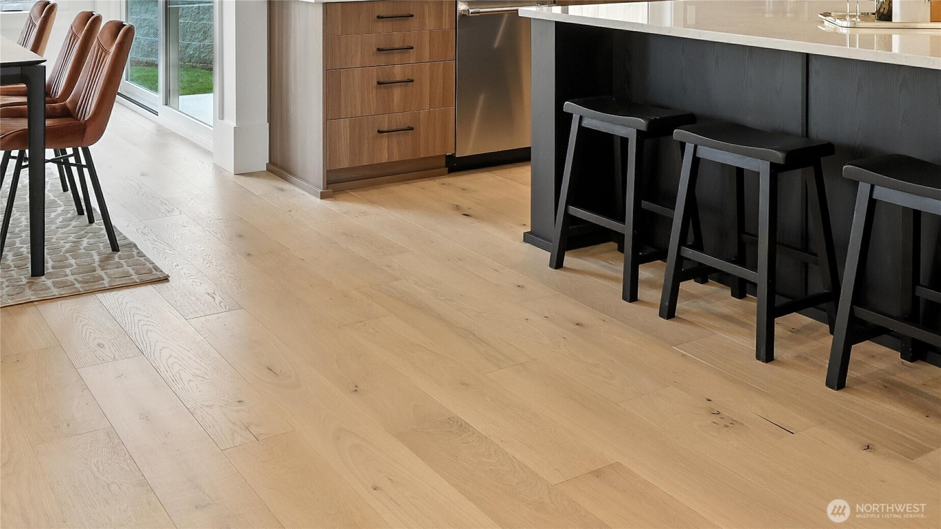 A striking centerpiece for daily life and entertaining alike. The chef’s kitchen pairs rich wood cabinetry with sleek black-and-brass accents, grounded by an oversized quartz island that doubles as a prep station and social hub. Pendant lighting adds modern warmth, while full-height windows draw natural light across the open-concept layout. The seamless flow into the great room highlights the home’s sense of space and connection — perfect for casual gatherings or elegant evenings by the fire.