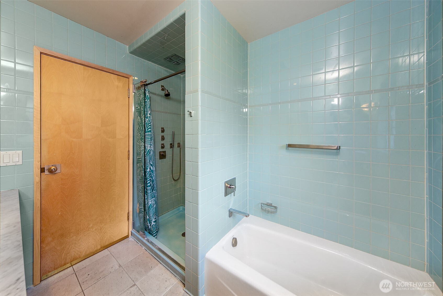 A calm, light-filled bathroom finished in soft blue tile features a walk-in shower with glass enclosure and a soaking tub set into a clean, modern surround. Warm wood accents and simple lines give the space a quiet, functional clarity.