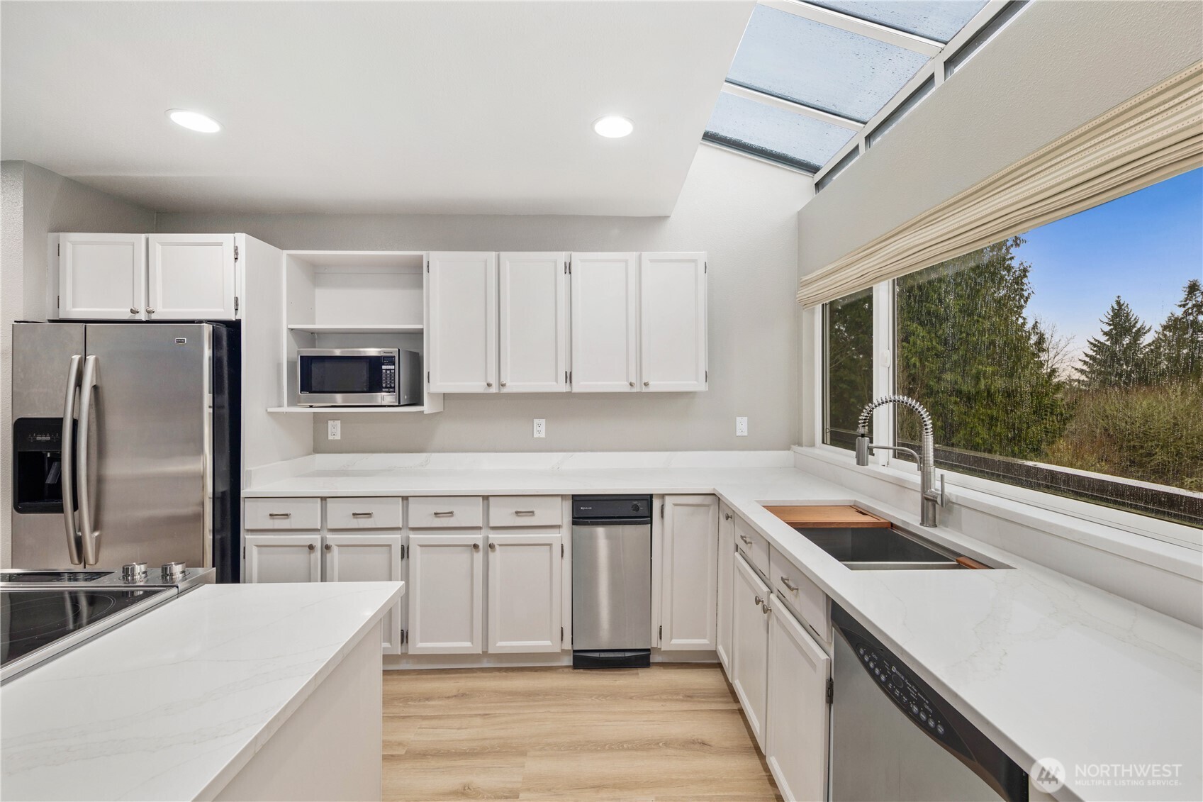 Open, sunlit kitchen with expansive island, seating, skylight, white cabinetry, stainless appliances, wide plank waterproof floors, and  an inviting entertaining layout.