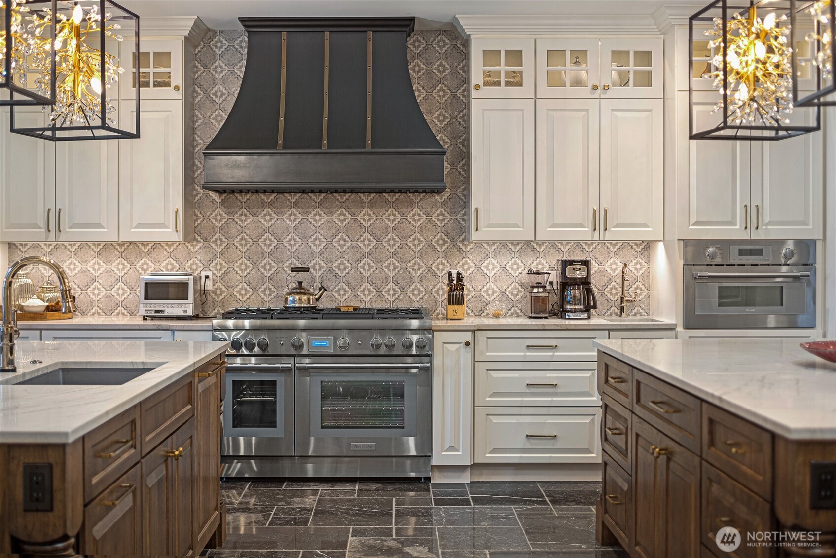 Professional stainless range centered beneath a custom metal hood and patterned tile backsplash, blending craftsmanship and luxury design.