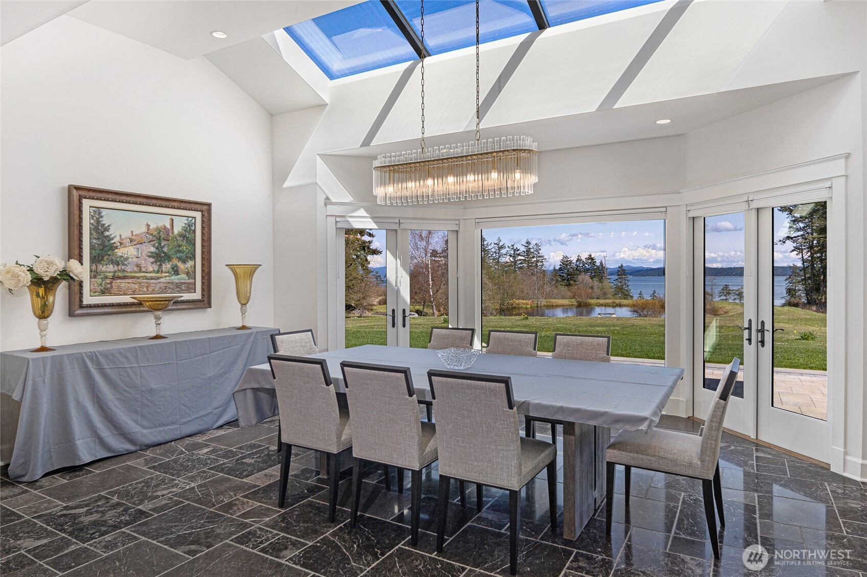 Light-filled formal dining room with skylight, modern chandelier, and expansive windows framing pond and water views.