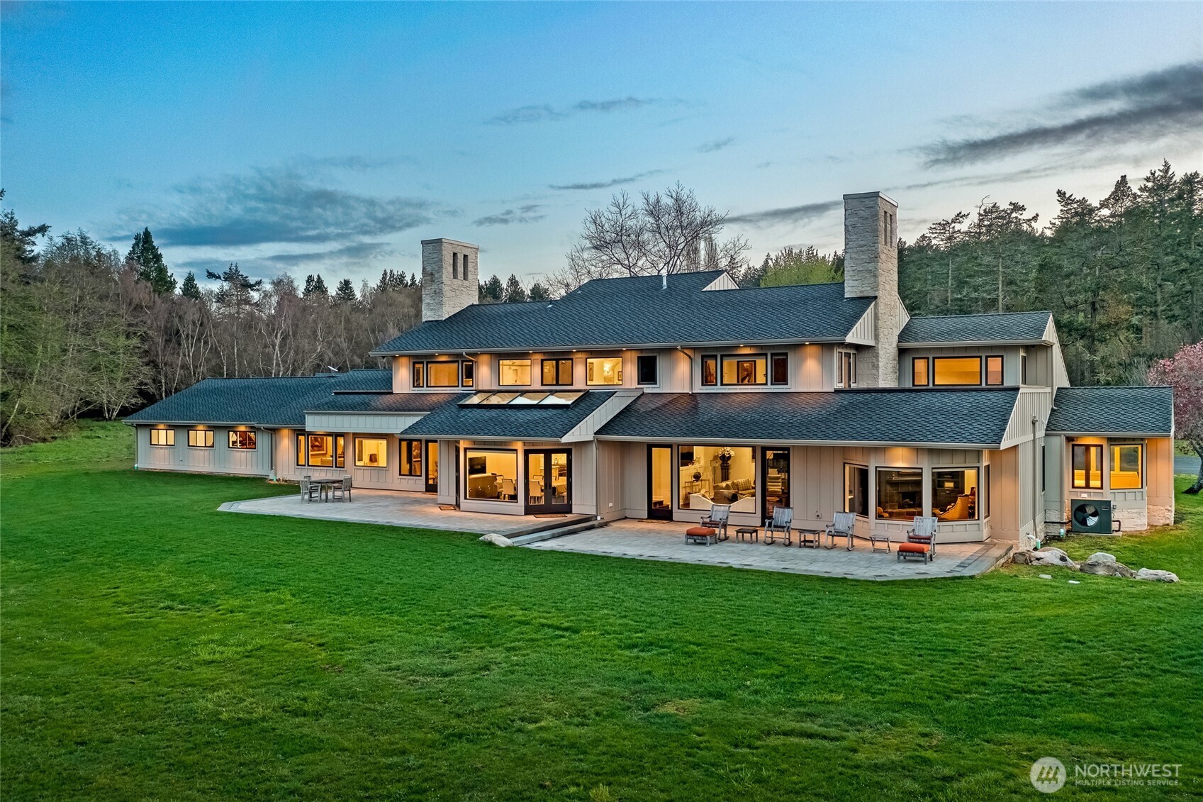 Twilight view of the estate showcasing warm interior glow, dual stone chimneys, and expansive patio spaces designed for elegant entertaining on San Juan Island.