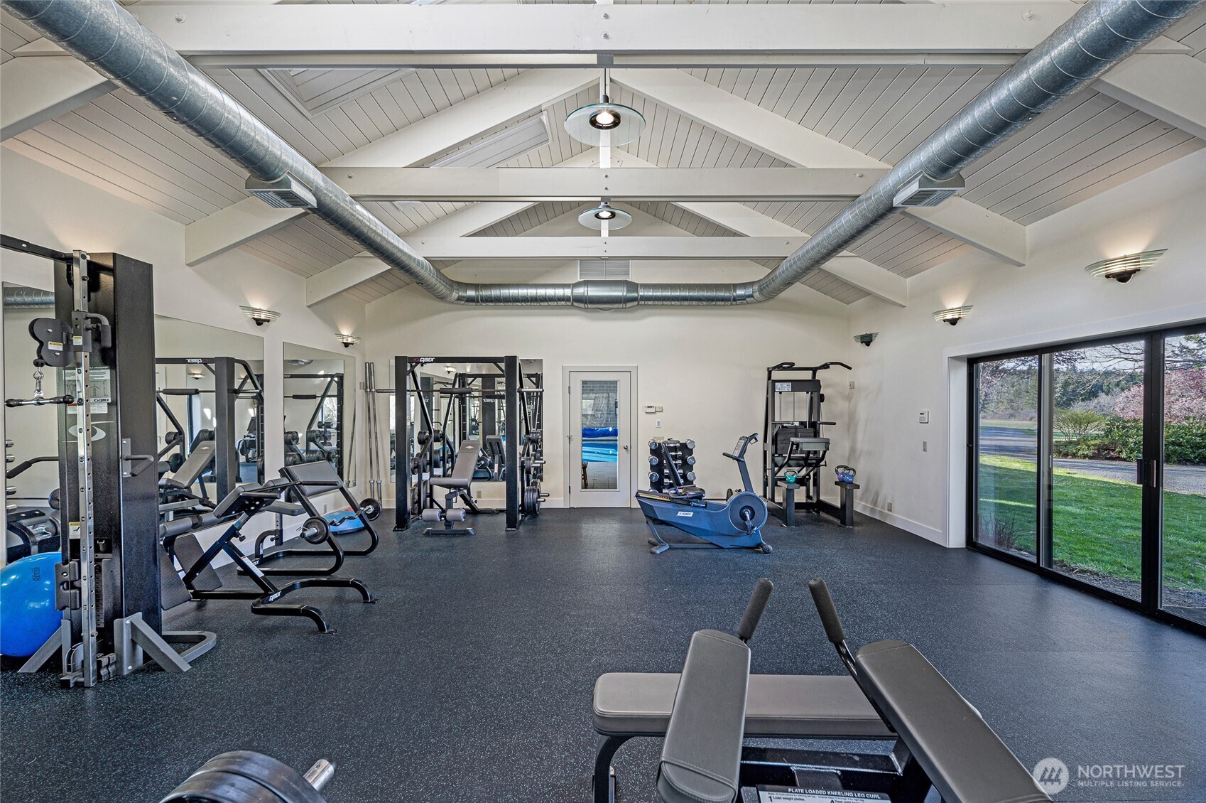 Private fitness studio with vaulted ceilings, exposed beams, full gym equipment, rubberized flooring, and sliding doors to exterior grounds.