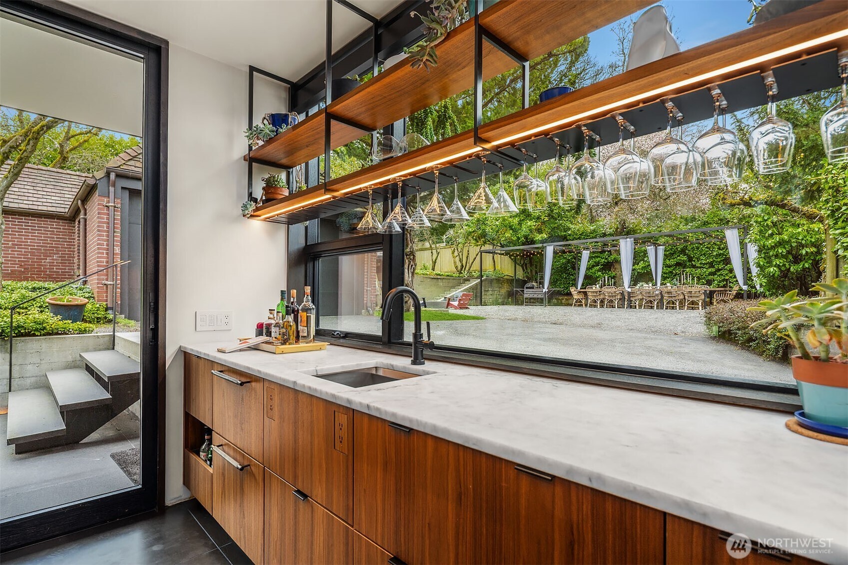 Steel and walnut shelves float on a cabinet to ceiling glass window; framing the outdoor Tuscan dining table