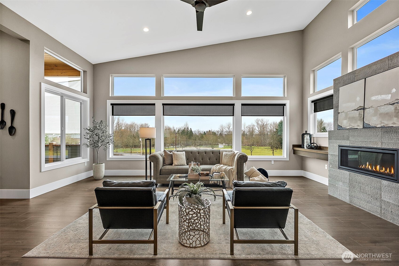 The living room features tall windows that frame open views while filling the space with natural light. A modern fireplace and open concept layout create a bright and welcoming living area designed for both entertaining and everyday comfort.