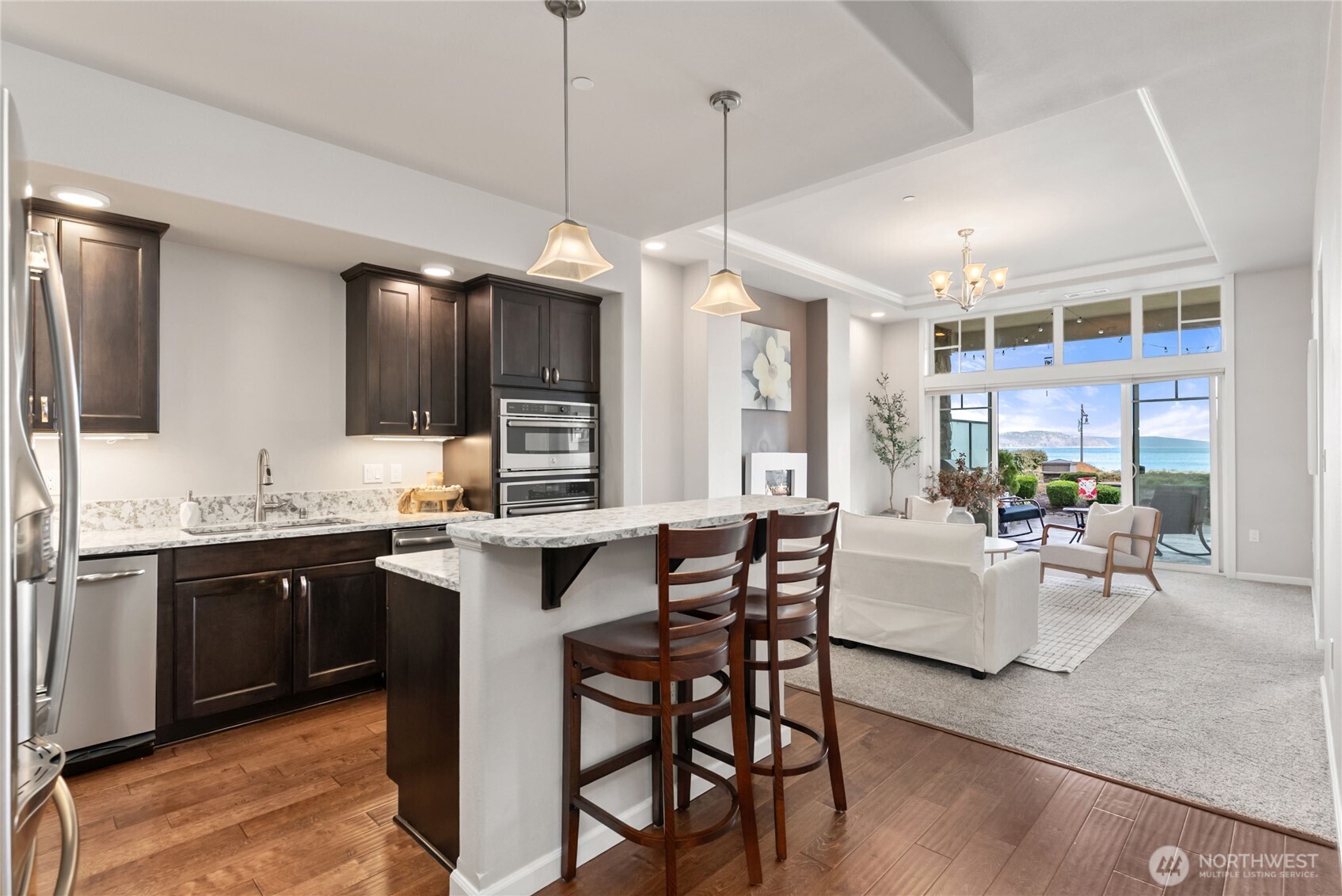 Open-concept kitchen and living space featuring rich cabinetry, granite countertops, stainless steel appliances, and a breakfast bar for casual dining. Pendant lighting and warm wood flooring create an inviting atmosphere that flows seamlessly into the living area and toward expansive waterfront views.