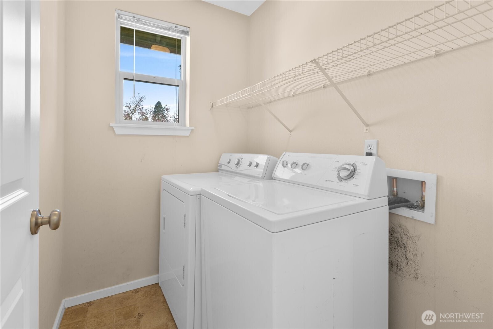 Laundry room with washer and dryer, wall-mounted wire shelf, and window providing natural light.