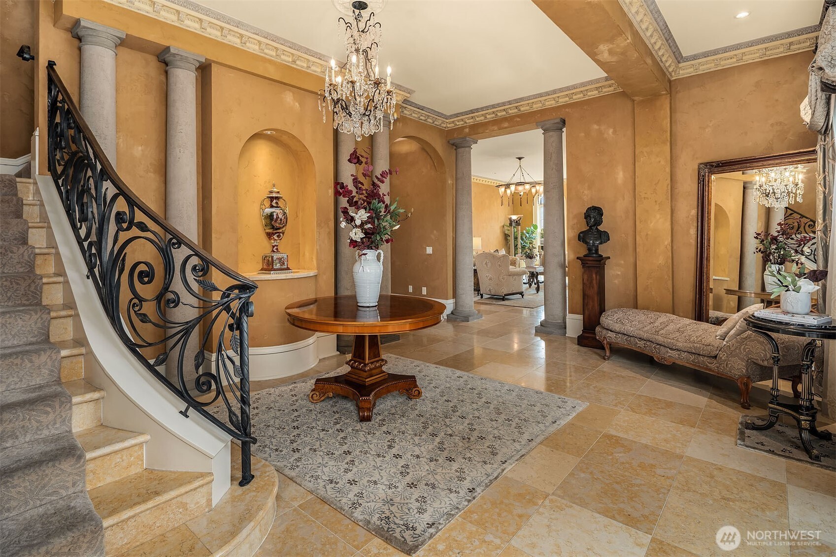 Step into Italian Renaissance-inspired interiors featuring Venetian plaster walls with ornate cornice moldings and Jerusalem gold marble floors throughout the main level. This grand foyer room showcases classical Roman columns, a sparkling crystal chandelier, and curved staircase with a custom wrought-iron railing featuring a Tuscan scrollwork design.