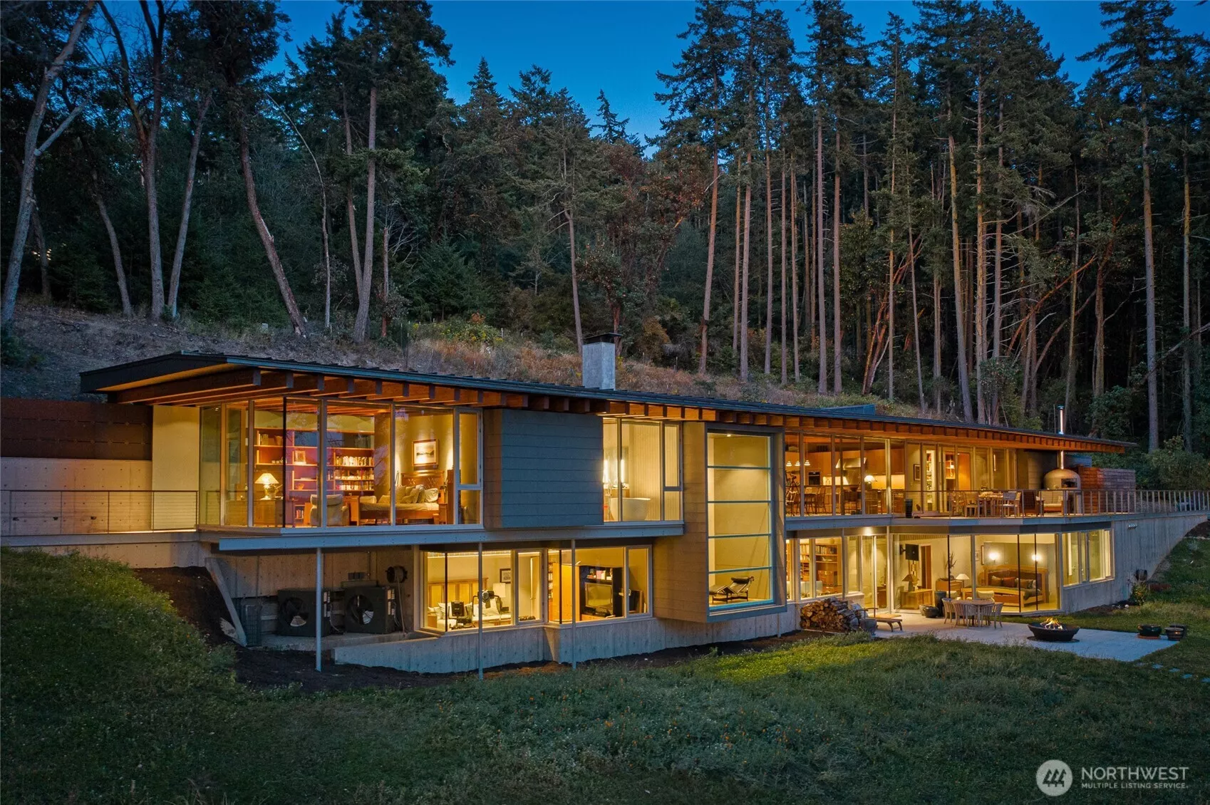 As evening falls, the residence glows warmly against the forested backdrop, highlighting the refined architecture and expansive glass walls that define this exceptional San Juan Island luxury waterfront estate.