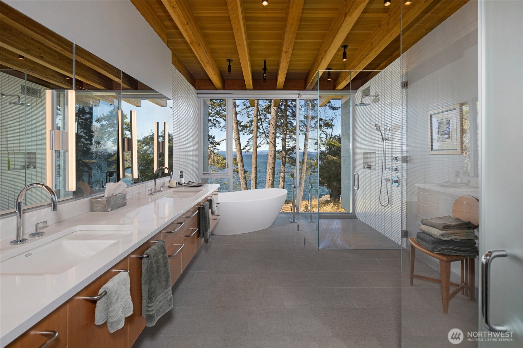 A spa-inspired primary bath features dual vanities, a freestanding soaking tub, and a glass-enclosed shower, all positioned to capture serene water views through floor-to-ceiling windows.