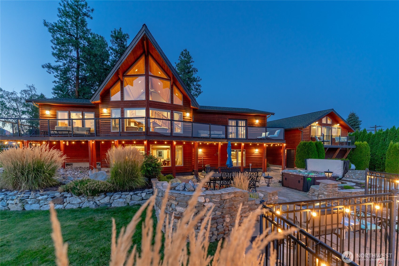 Twilight view of home w/ oversized windows, view decks, outdoor living area w/ pool, hot tub, basketball court, dining and lounge areas and detached carriage house.