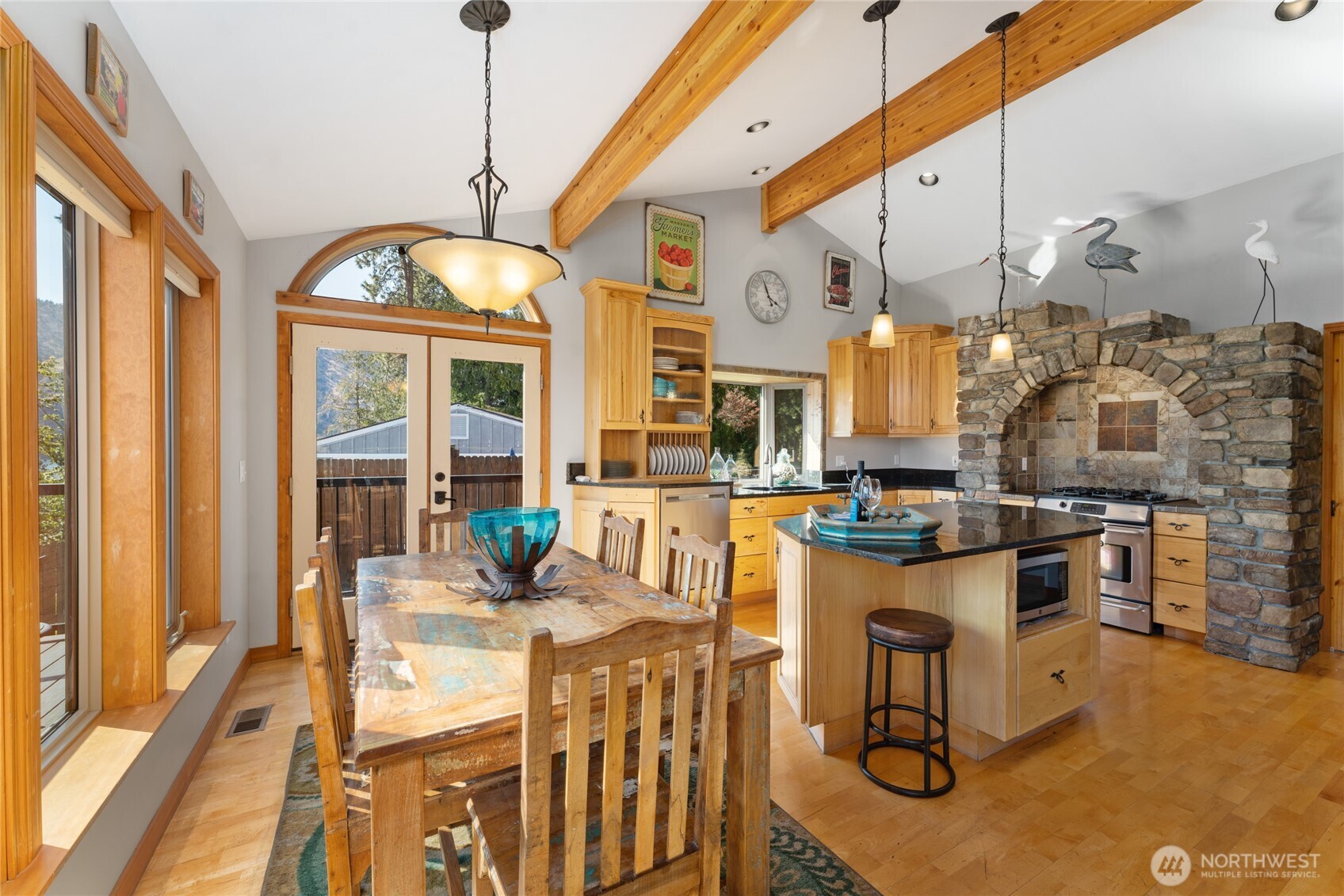 View of dining room and kitchen w/ hardwood floors, stainless steel appliances, quartz countertops, wood beams,  stone surround stove and oven, large windows with views of Lake Chelan and French doors leading to the outdoor living area w/ pool, hot tub, dining and lounge area.