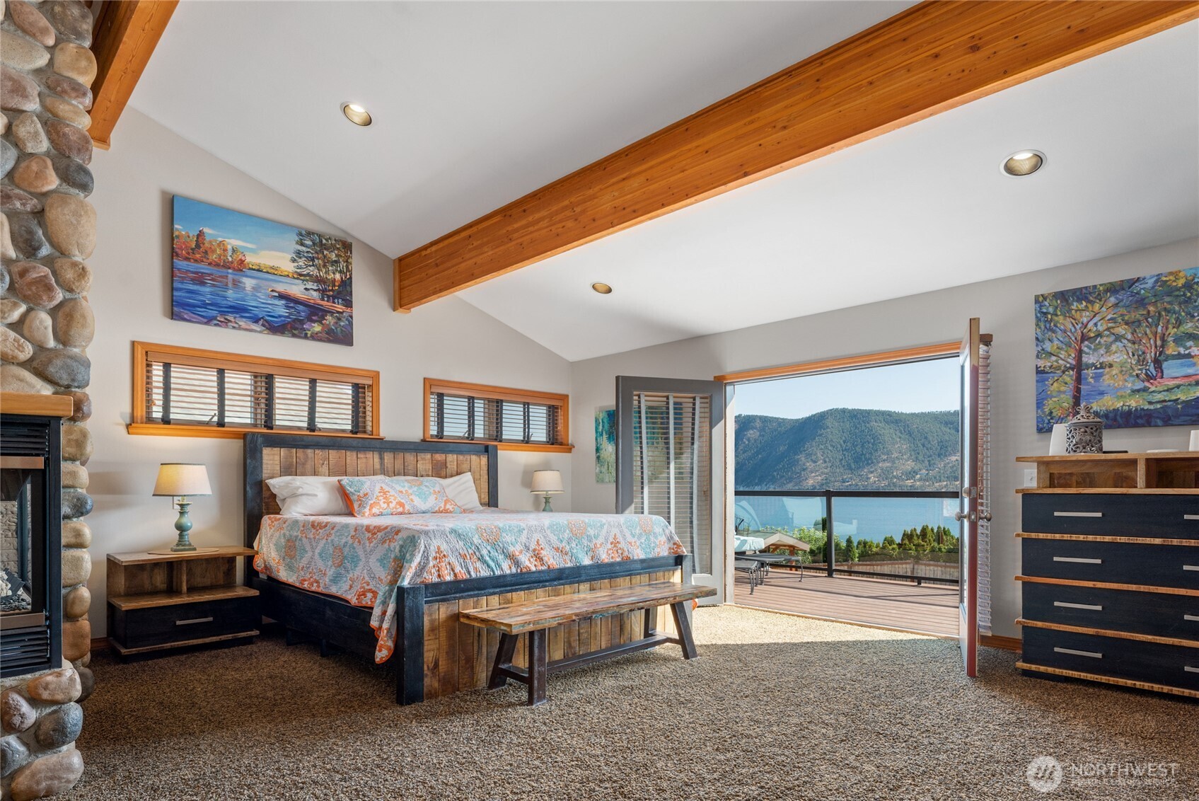 View of main level Primary Bedroom suite w/ stone fireplace and French Doors leading to the lake view deck seating area w/ glass railings.