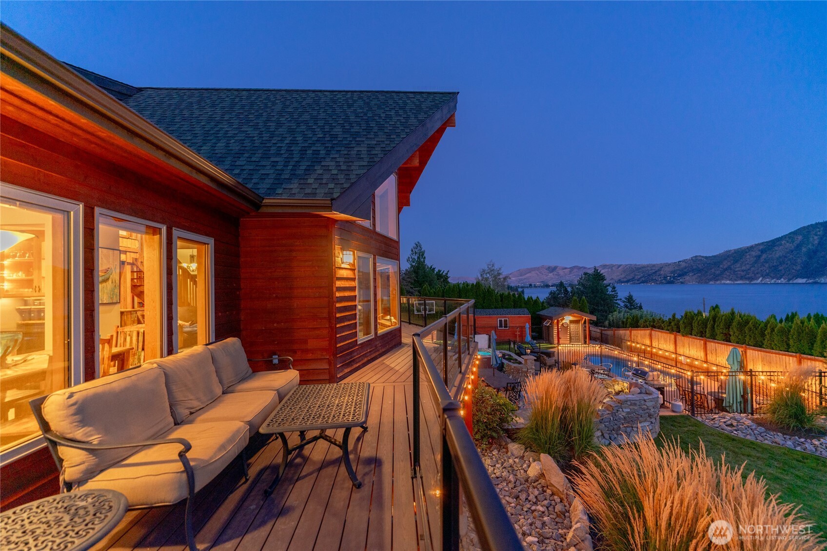 Twilight view of home w/ oversized windows, view decks, outdoor living area w/ pool, hot tub, basketball court, dining and lounge areas and landscaped grounds overlooking Lake Chelan.