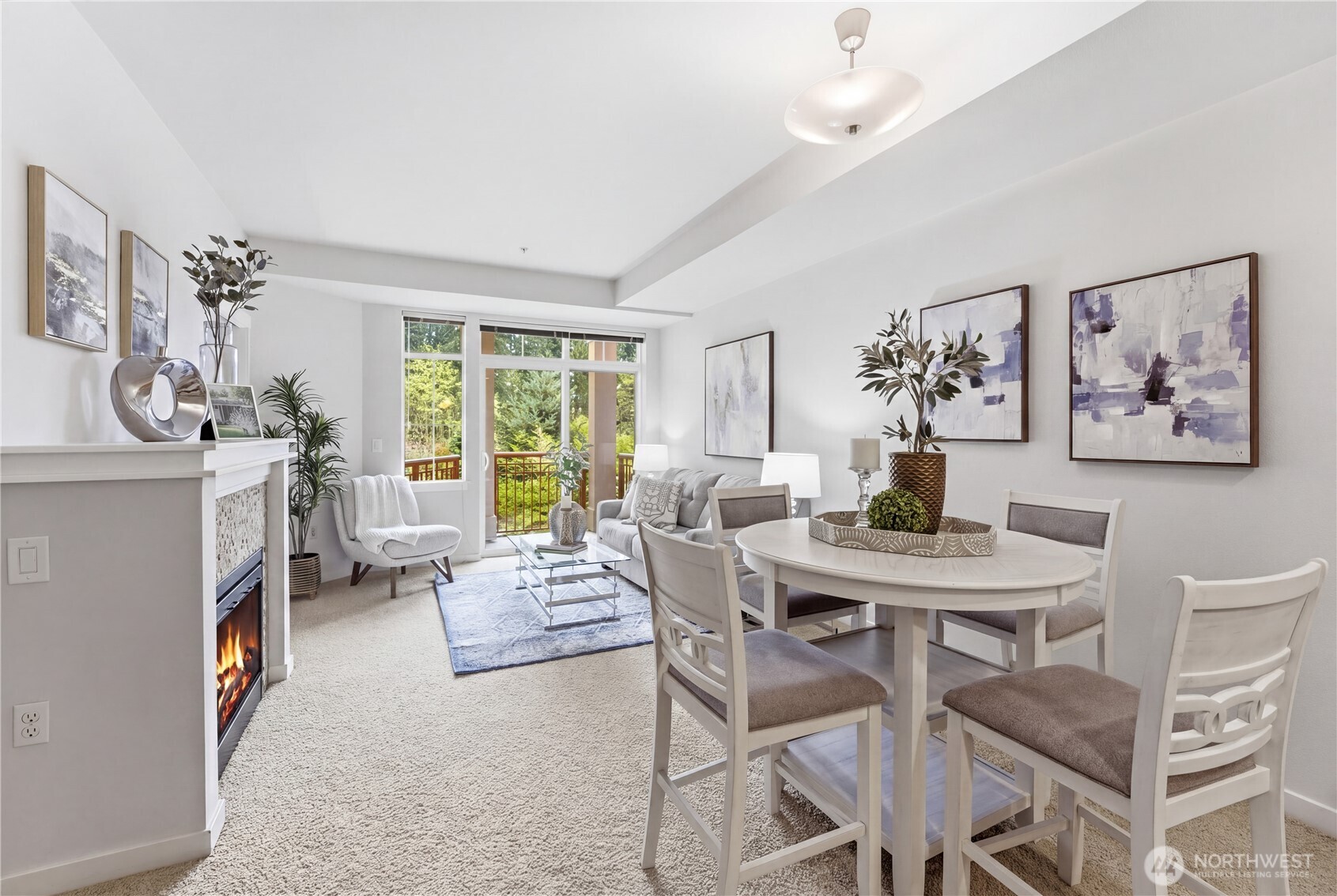 Spacious living and dining area with a fireplace, coordinated artwork, and large windows that fill the room with light and views of the trees