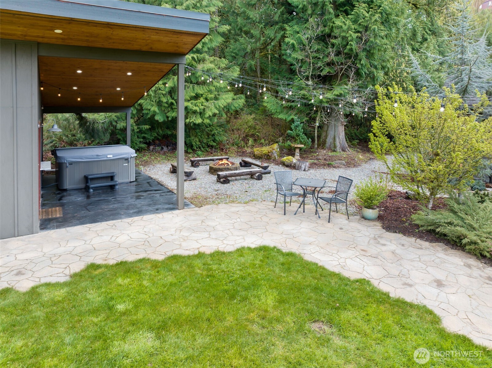 Perfect for outdoor gatherings, adjacent to the cabana, this inviting outdoor retreat offers a covered hot tub area, a cozy fire pit surrounded by rustic seating, and a patio for casual conversation—all set beneath string lights and surrounded by mature evergreens