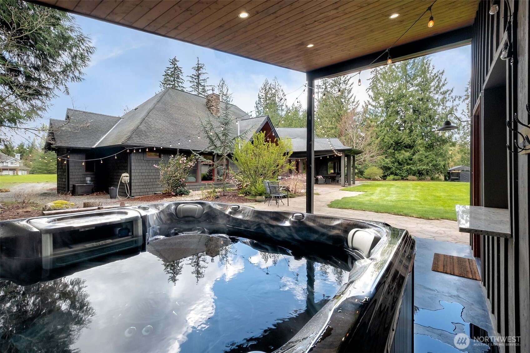 From the covered hot tub, enjoy a serene view across the manicured lawn to the beautifully designed home and outdoor living spaces—offering a peaceful, resort-like setting surrounded by mature evergreens.