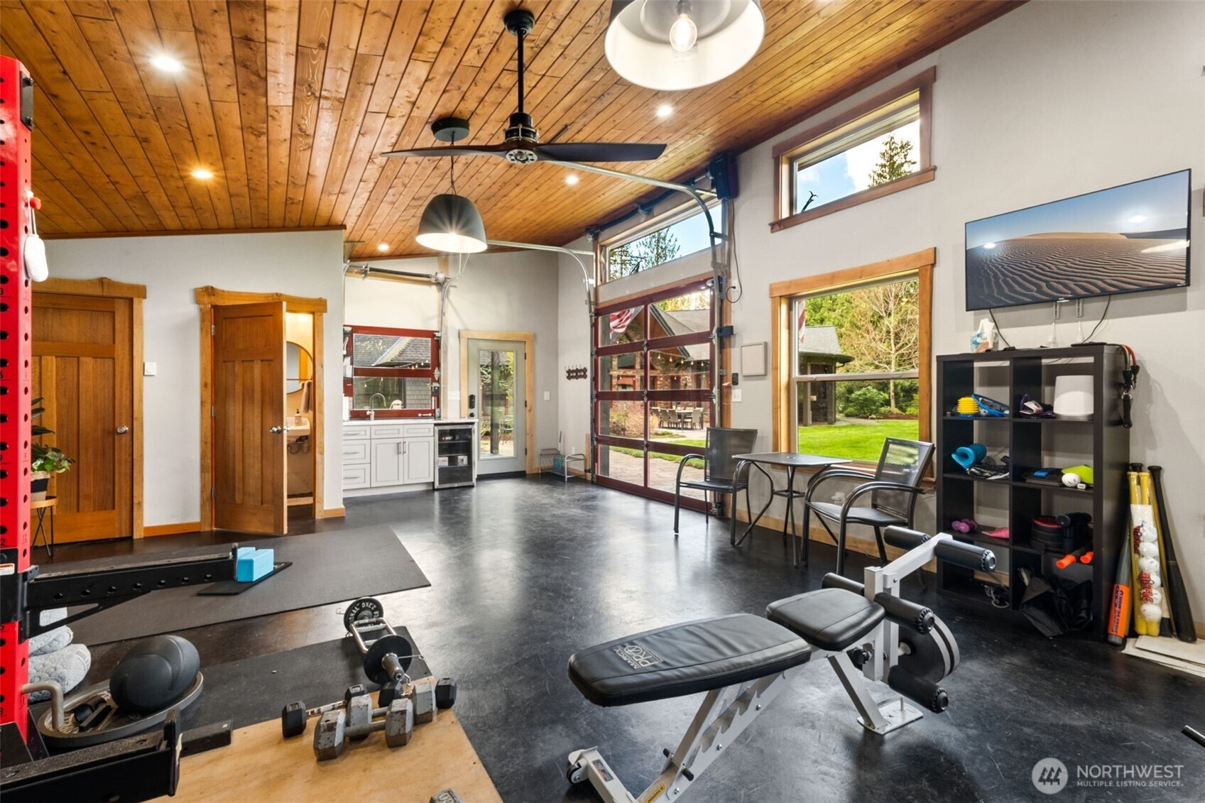Currently used as an exercise space, the cabana’s interior showcases vaulted tongue-and-groove wood ceilings, contemporary lighting, and ceiling fans for comfort. Clerestory windows bring in additional natural light, while the glass garage door opens wide to the outdoors—perfect for gatherings, workouts, or studio use.