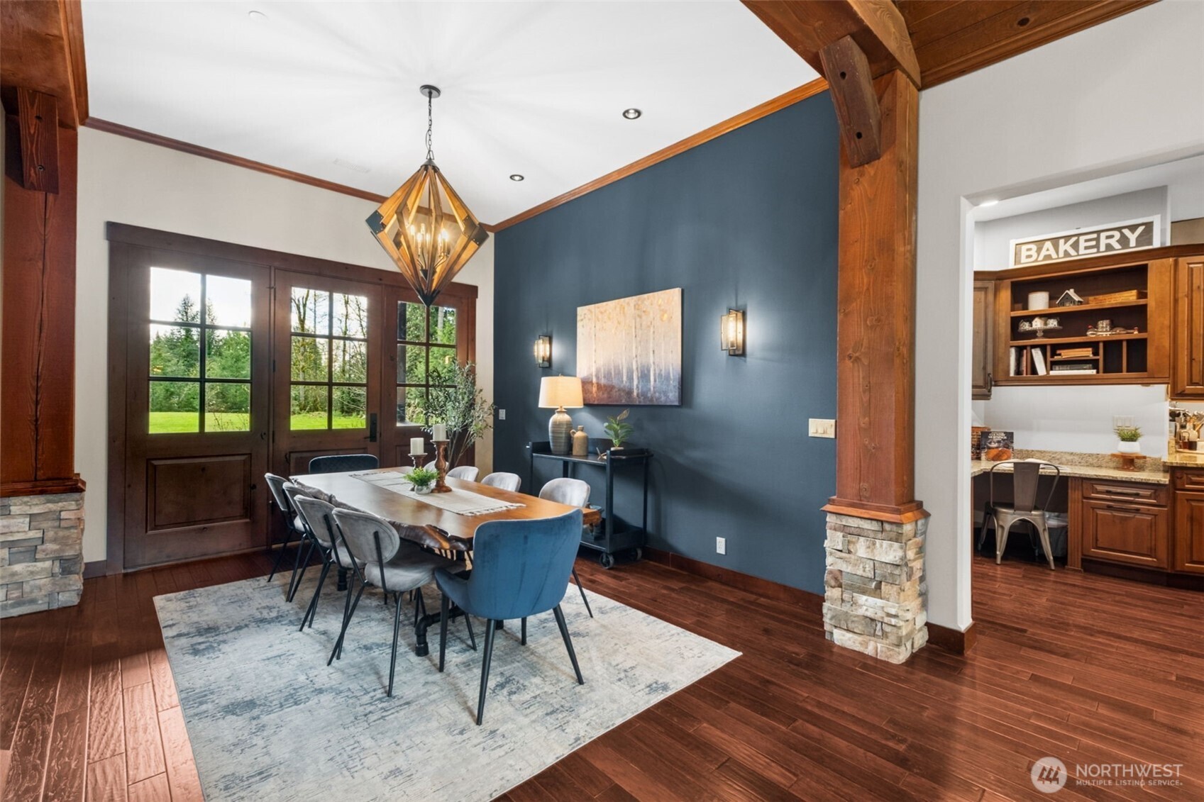 A richly painted accent wall adds depth and contrast to the dining area, complemented by warm wood flooring and exposed timber details. The statement chandelier anchors the space, while French doors flood the room with natural light and connect seamlessly to the outdoors.