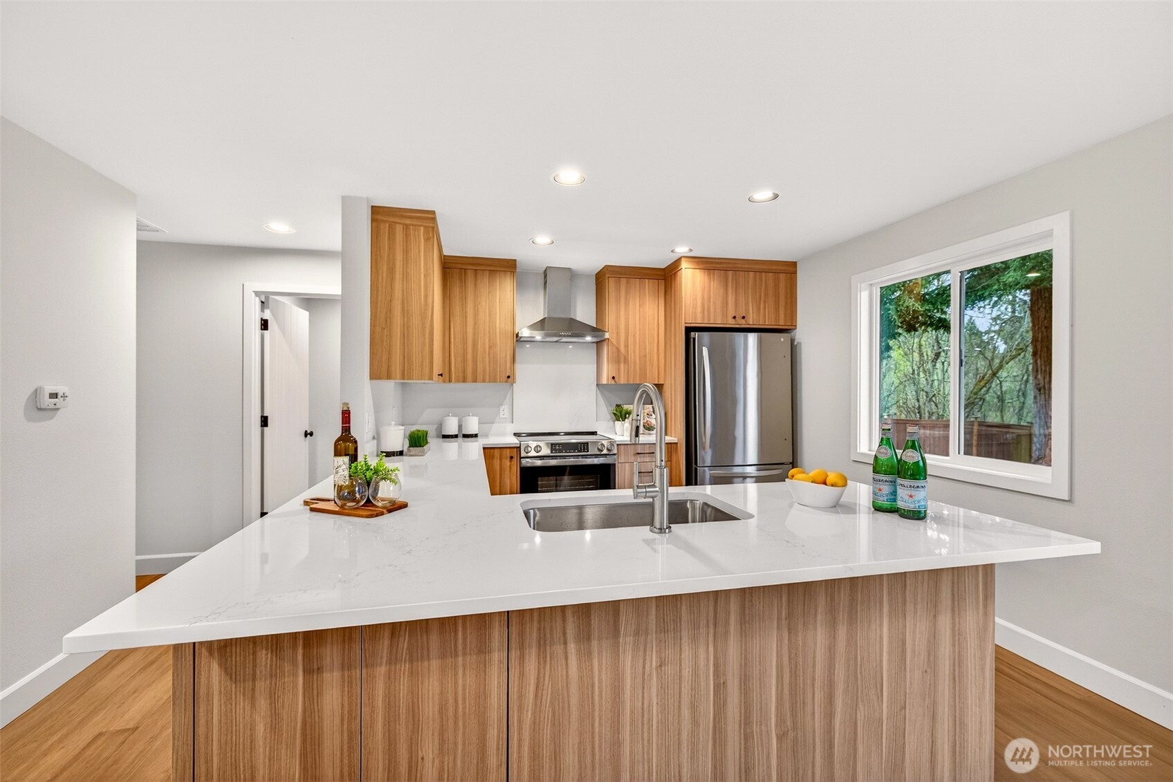 Expansive quartz countertops and a deep undermount sink anchor the kitchen peninsula, creating ideal prep space for everyday cooking and weekend entertaining alike. A picture window above the sink frames the wooded greenbelt — making even routine moments in this kitchen feel connected to nature.