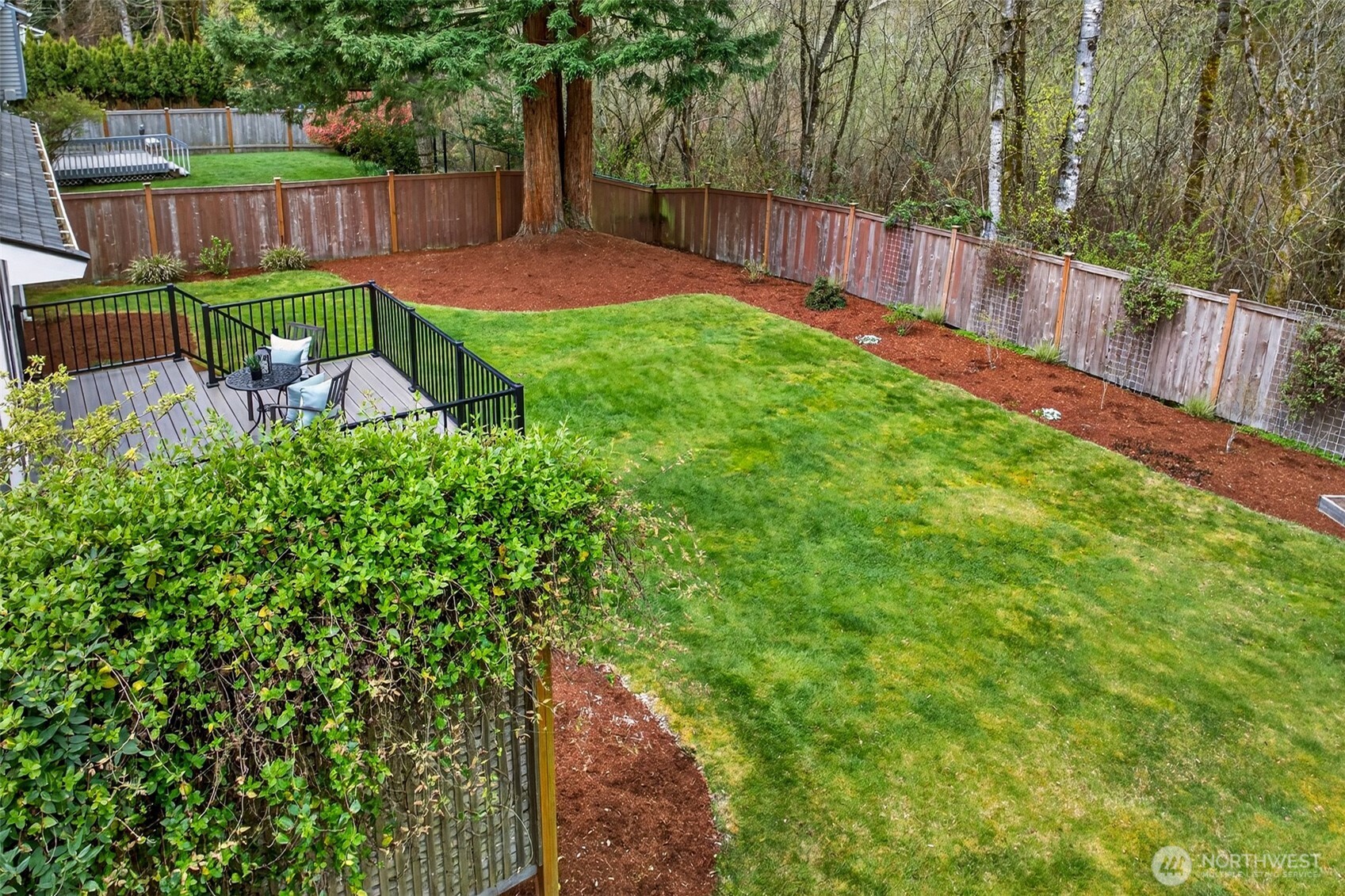 This elevated perspective reveals just how generously sized and thoughtfully landscaped the rear yard truly is — a sweeping fenced lawn with mature tree anchors, mulched borders, and a composite deck positioned for both privacy and connection to the natural greenbelt beyond. It's a complete outdoor environment that elevates the entire living experience of this home.