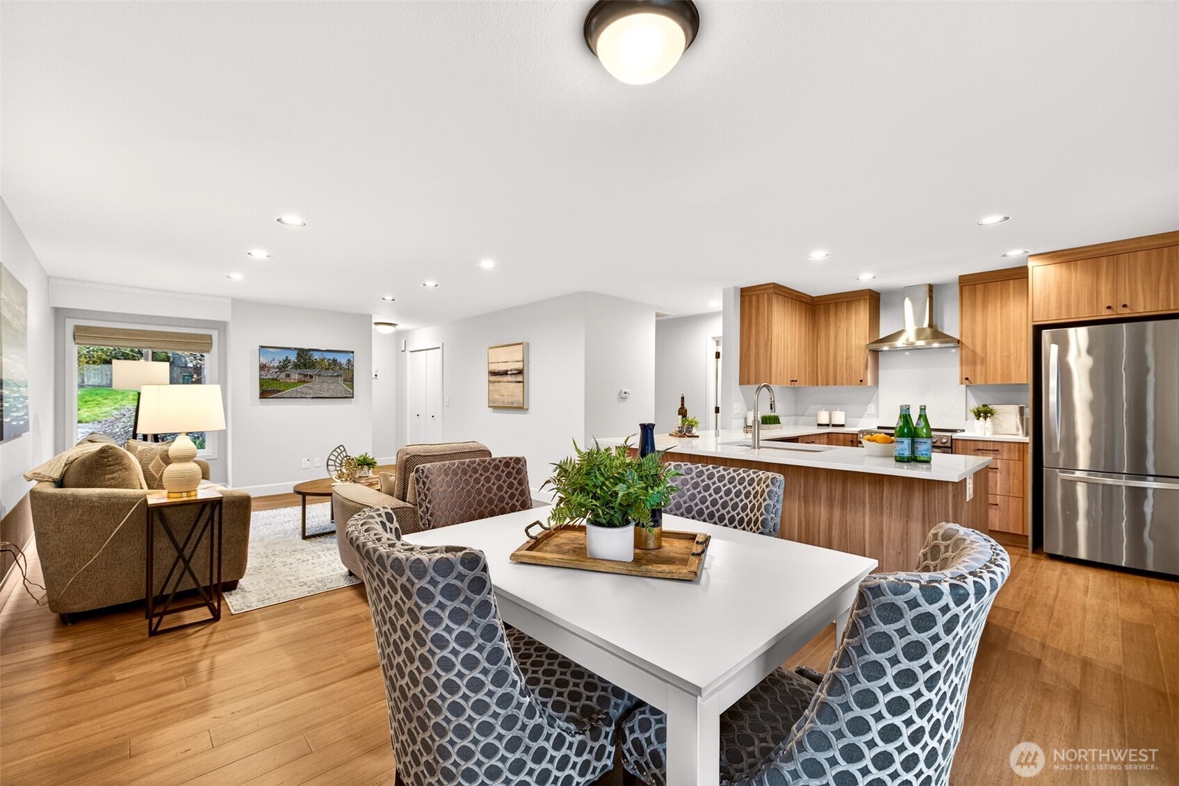 This angle captures the full depth of the open-concept main level — from the updated kitchen with stainless refrigerator and custom walnut cabinetry all the way through to the living area — demonstrating just how livable and connected the floor plan truly is. Updated hardwood flooring ties the entire space together with warmth and continuity.
