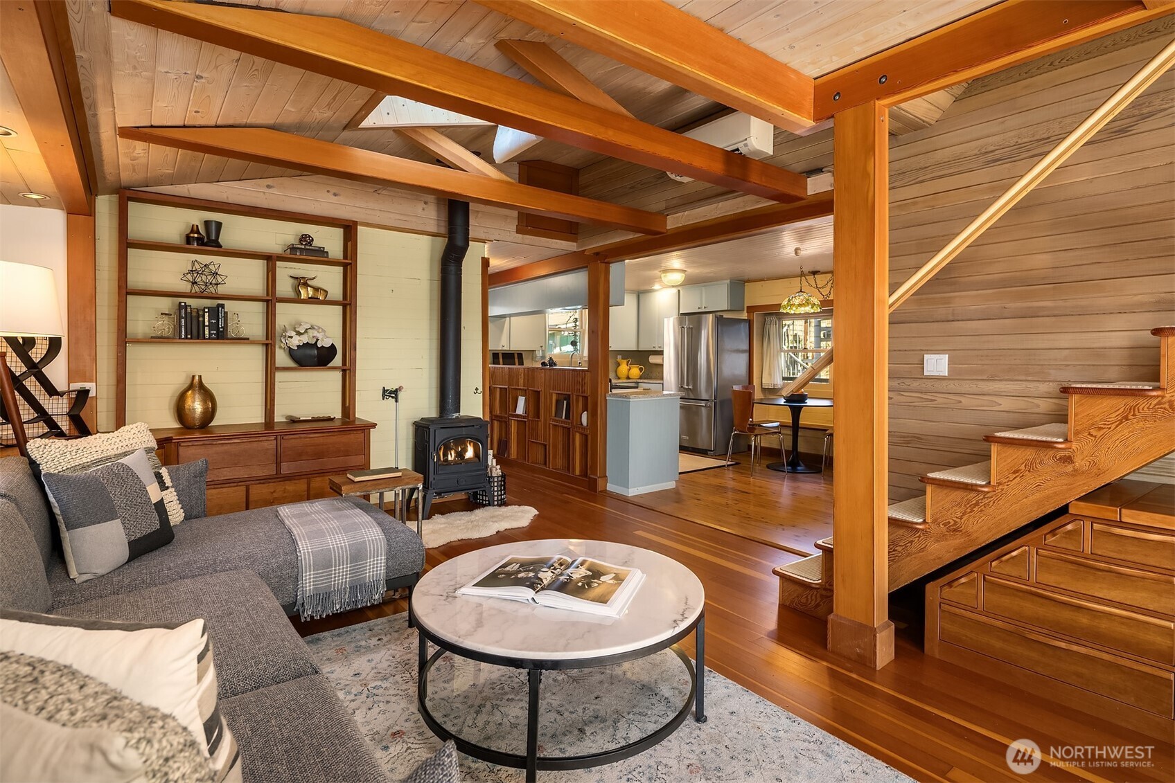 The living room looking toward the kitchen — wood stove, built-in shelving, vaulted beams, and a sectional sofa that invites you to settle in and stay a while.