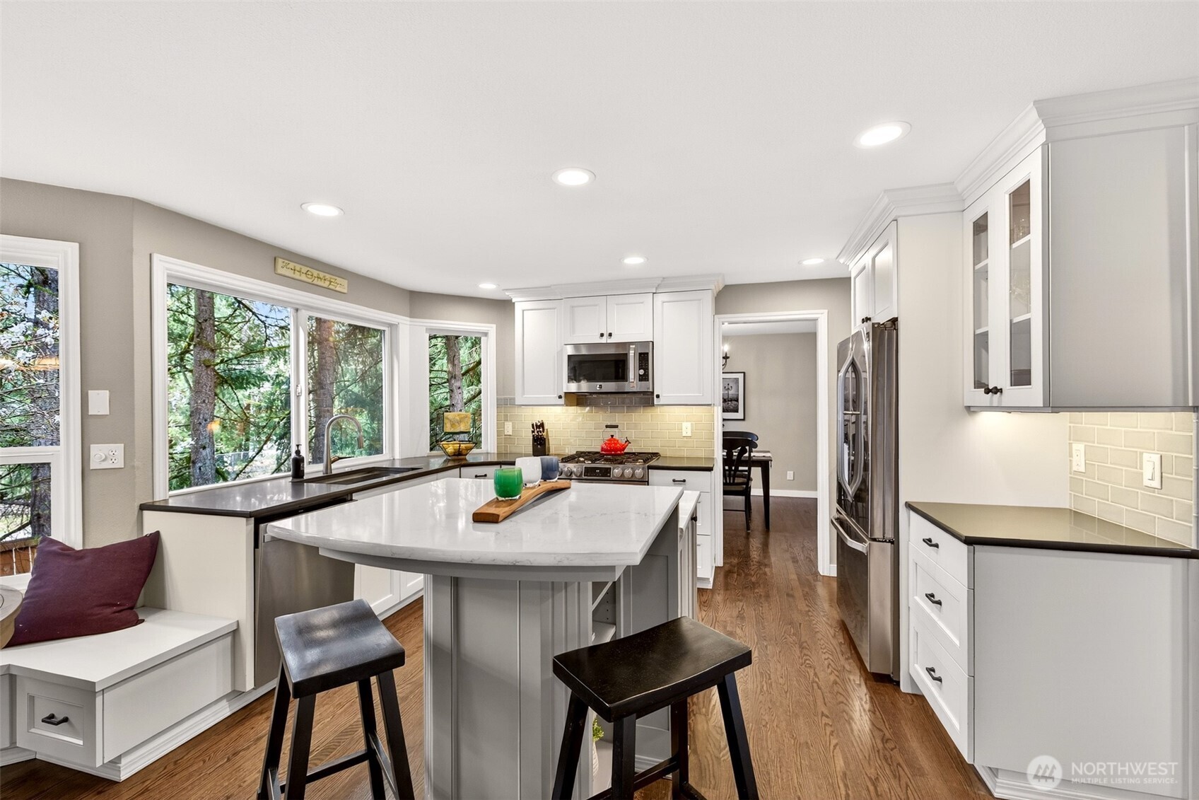 A well-designed kitchen layout with a quartz-topped island, built-in breakfast nook, wraparound windows overlooking the evergreens, and a seamless connection to the family room — the kind of space where mornings become a pleasure.