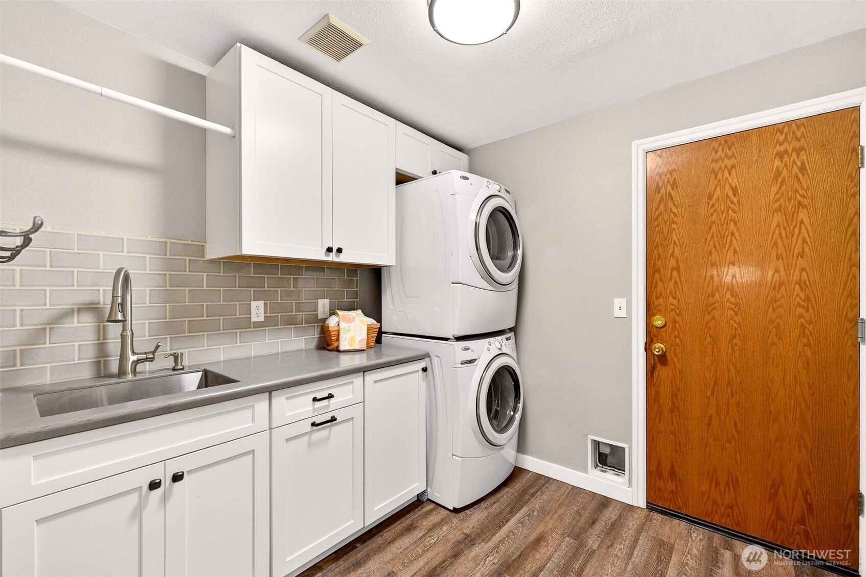 The updated laundry room featuring white shaker cabinetry, subway tile backsplash, utility sink, stacked washer/dryer, and direct garage access for added everyday convenience.