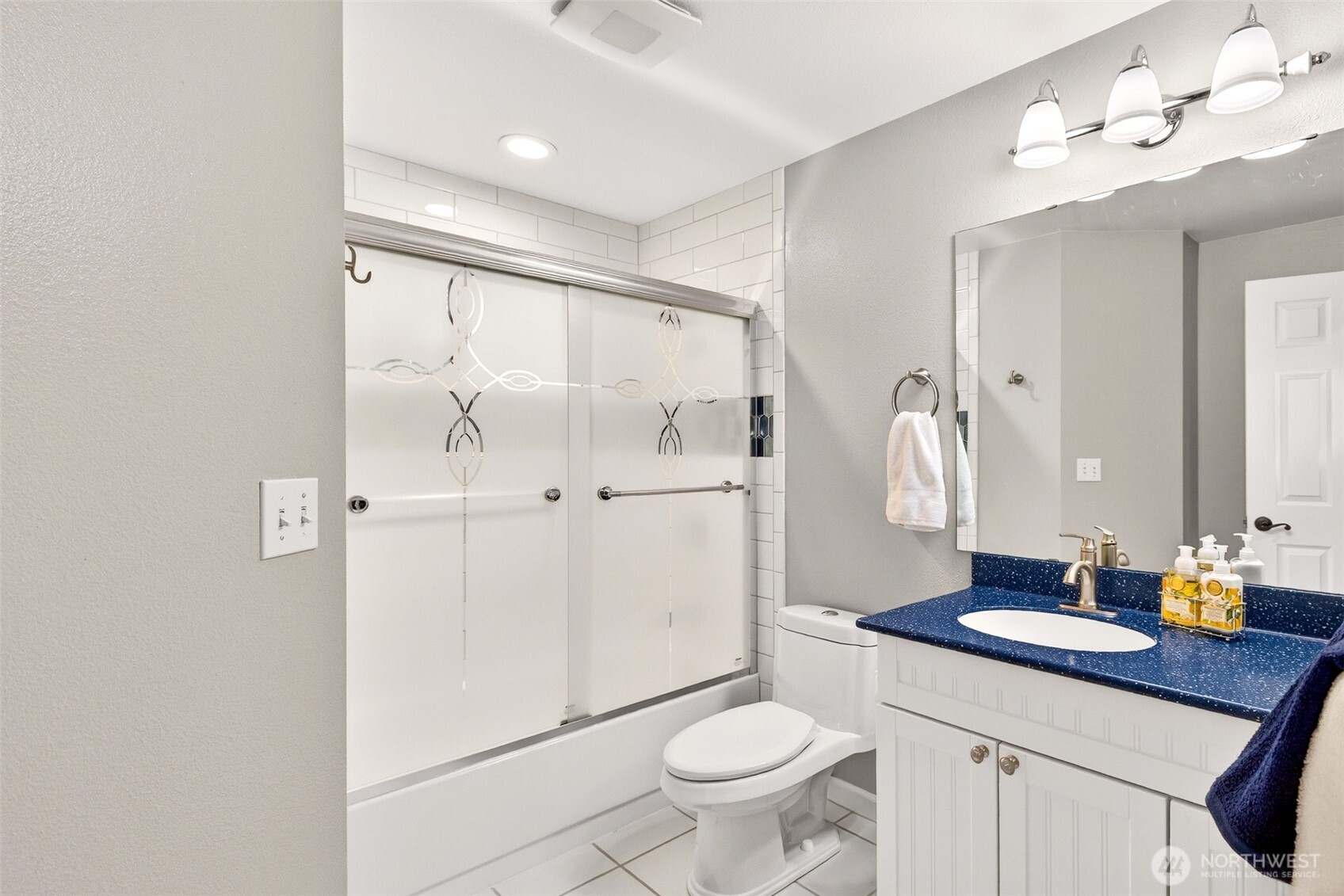 The full hall bath serves the secondary bedrooms with a clean, functional layout — white cabinetry, a bold blue seamless countertop, subway tile tub surround, and decorative sliding glass doors add personality and polish.