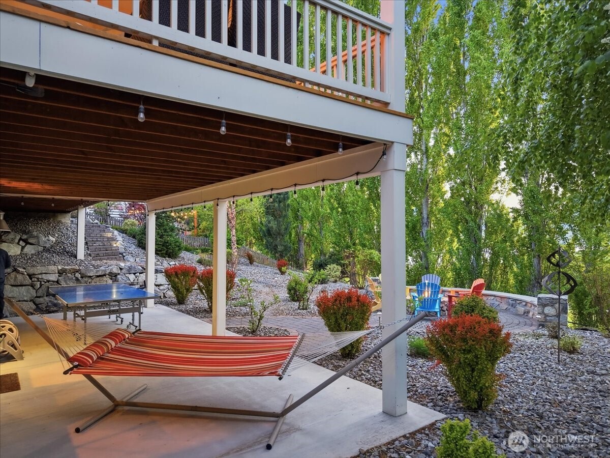 Exterior view of lower level outdoor living area with hammock, ping pong table and custom built-in stone fireplace with Adirondack chairs overlooking Lake Chelan.