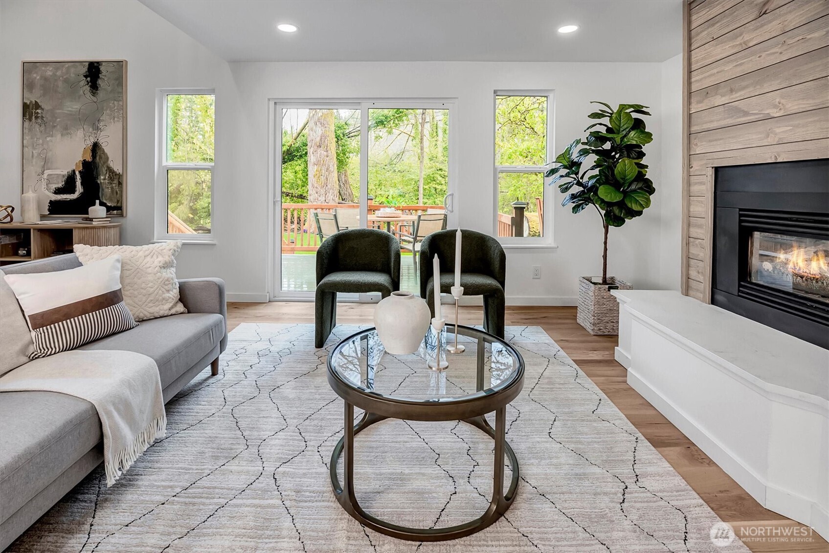 Living room featuring a wood-clad fireplace, laminate hardwood floors, and sliding glass doors opening to a deck with serene creek and tree-lined views.