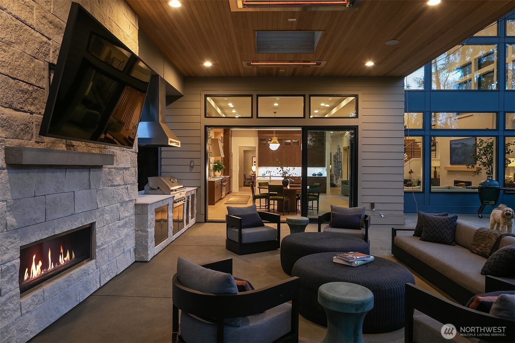 The flow from covered terrace to gourmet kitchen is the quiet genius of this floor plan. Fireplace glowing outside, kitchen lit warm within — the line between inside and out disappears entirely.