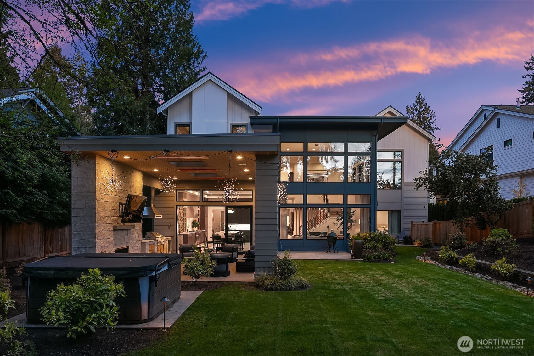 The full rear elevation at golden hour — covered terrace with outdoor fireplace and BBQ, floor-to-ceiling windows revealing the great room and kitchen beyond, and a pink-streaked sky above it all. Indoor-outdoor living without the compromise.