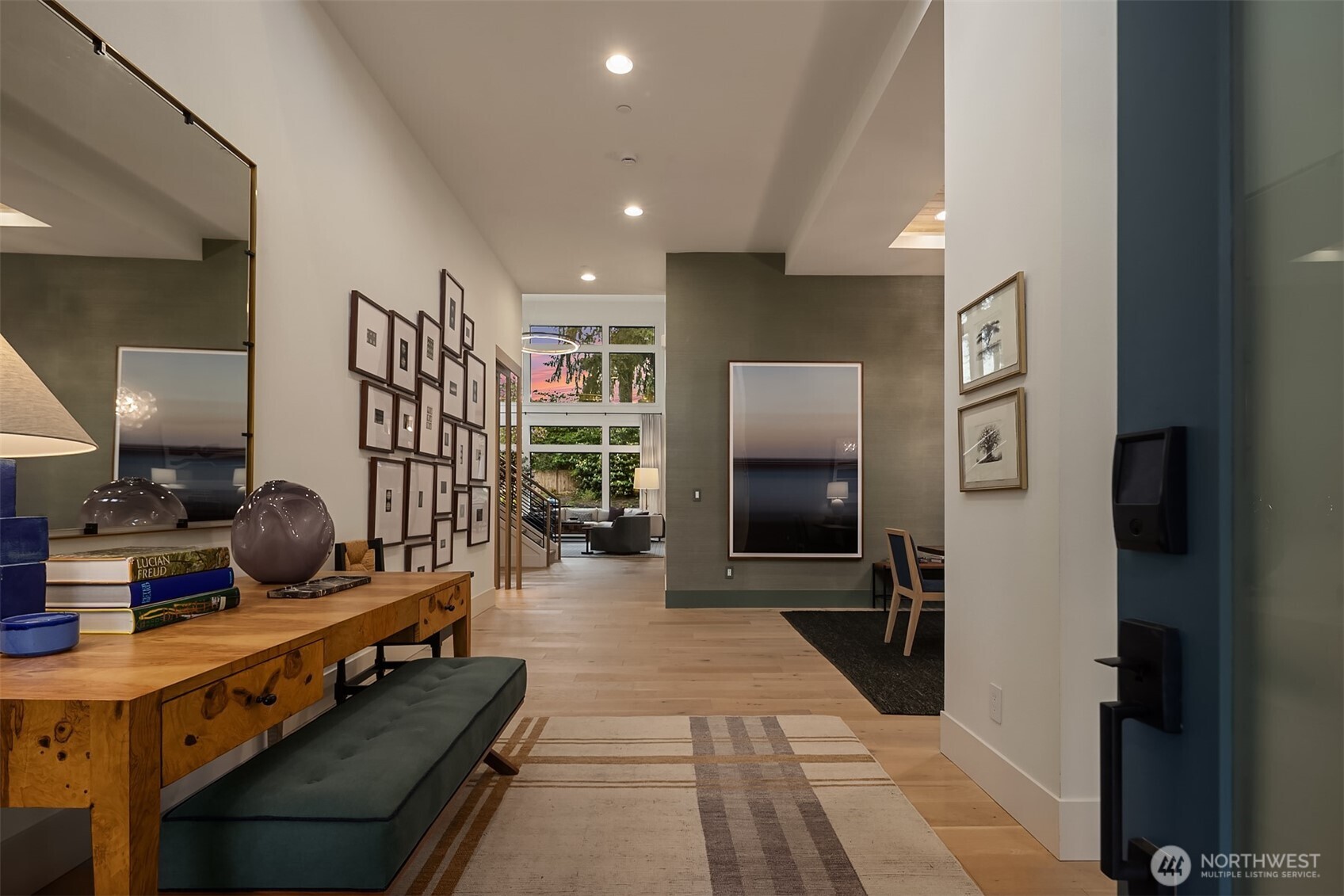 A generous entry that sets the tone before you reach the great room — wide-plank engineered white oak floors, soaring ceilings, and a pink-streaked sky visible straight through to the two-story window wall beyond.
