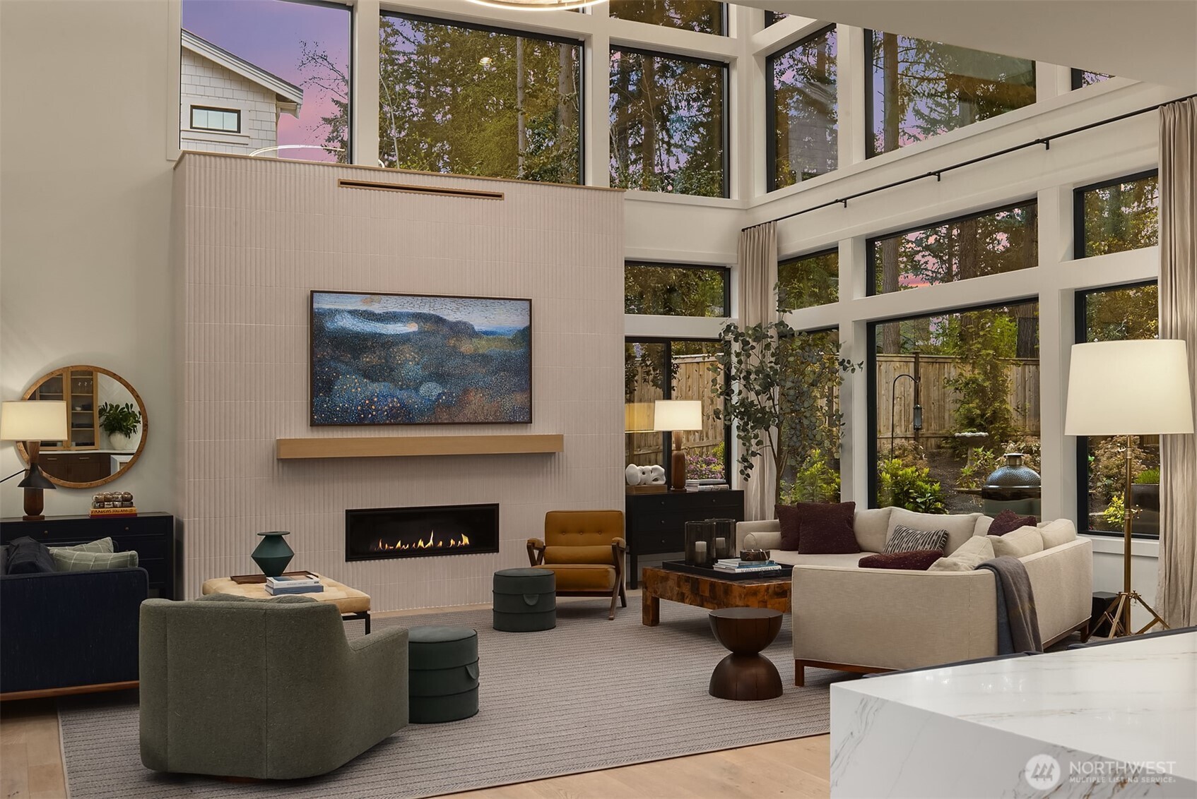 The great room from the kitchen's edge — fluted tile fireplace wall, two-story window wall, and a garden beyond the glass. The view you get every time you pour your morning coffee.