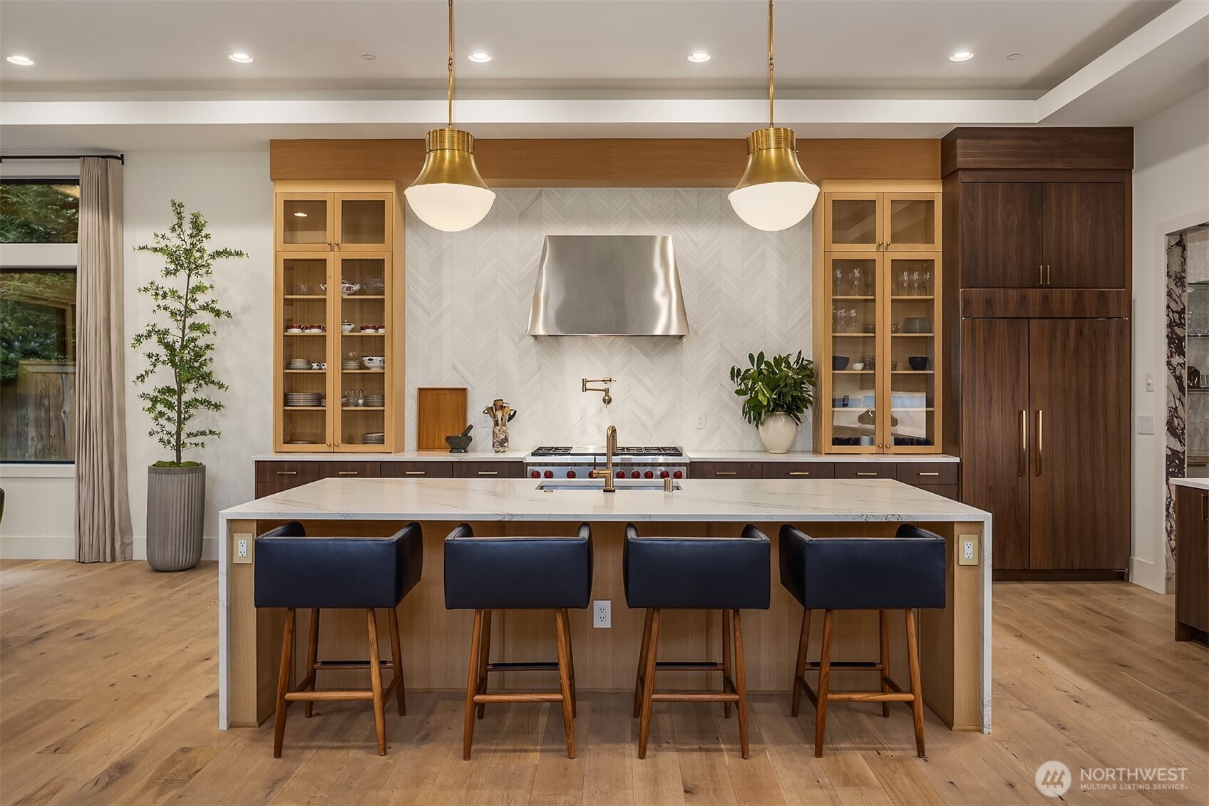 A waterfall-edge island, hand-selected honed Chevron Dolomite backsplash, stainless hood, and gold pendants overhead. The kitchen that makes you want to cook something worth talking about.