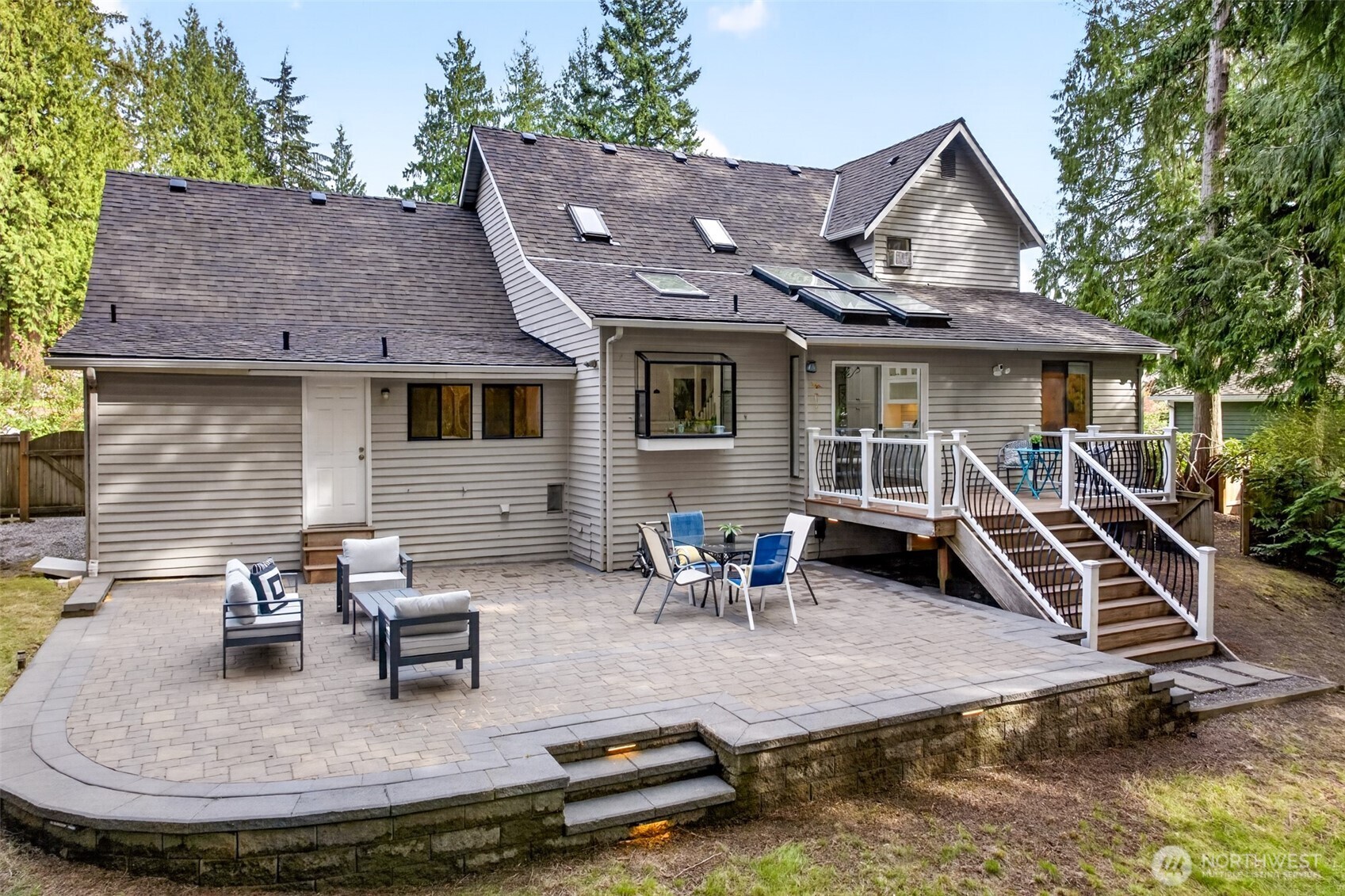 The full rear elevation showcases the freshly finished deck, expansive new paver patio with retaining wall and integrated lighting, and skylight-studded roofline tucked into a wooded setting.