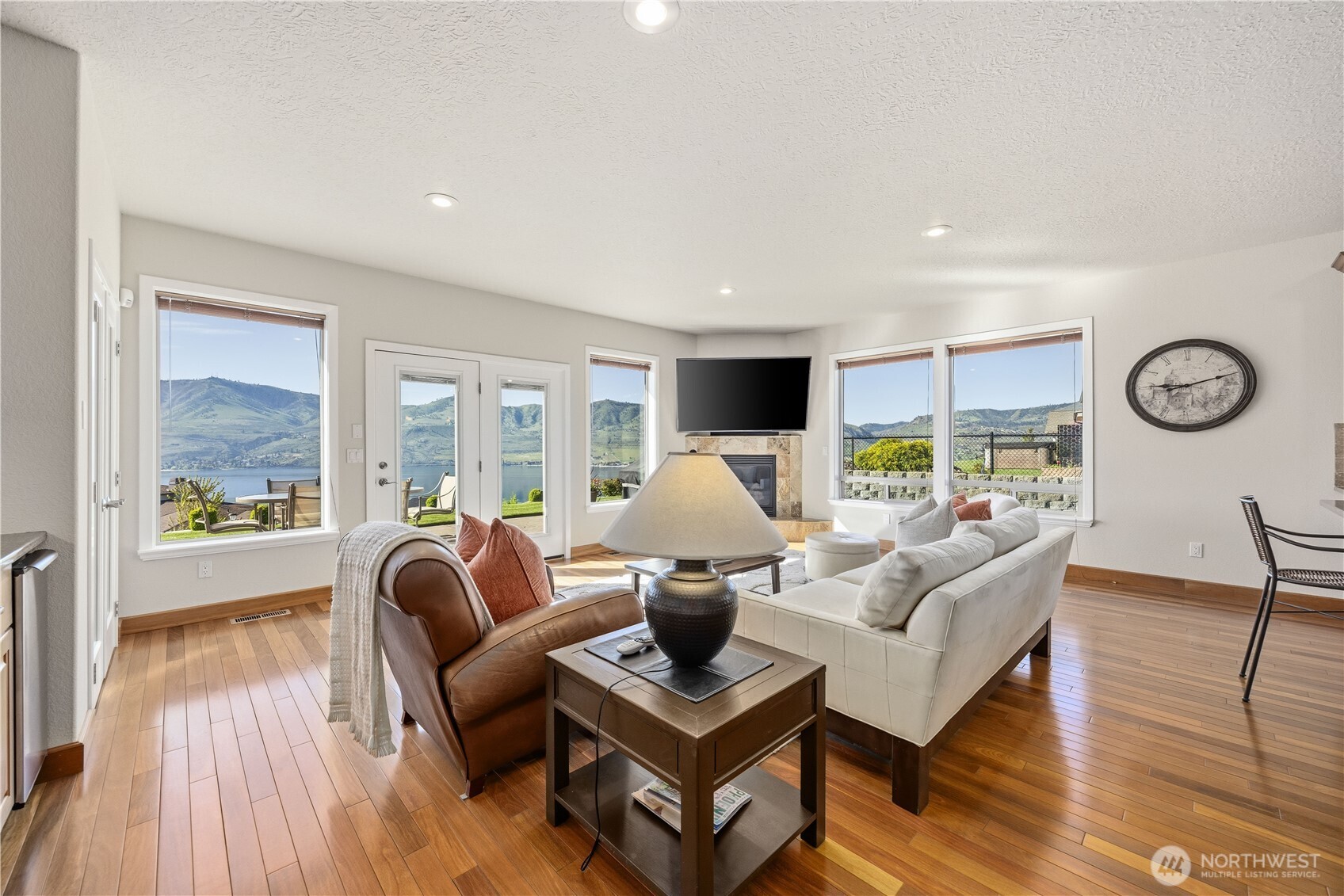 Bright and inviting living space with warm hardwood floors and expansive windows showcasing stunning Lake Chelan and mountain views. The open layout flows effortlessly to outdoor living through glass doors, while a cozy fireplace creates the perfect focal point for relaxing or entertaining in this scenic setting.