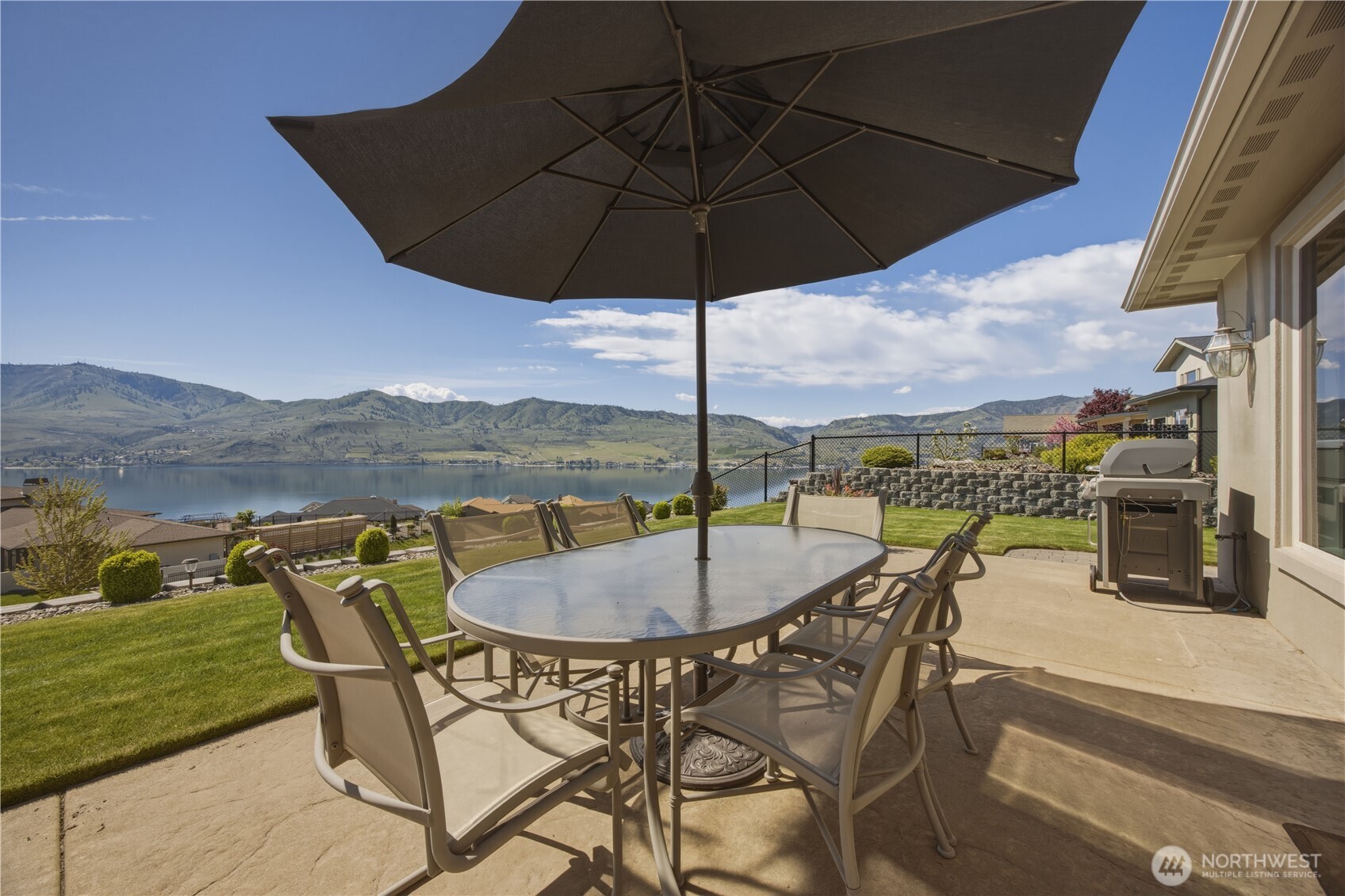Inviting outdoor dining area on the expansive patio, complete with shade umbrella and seating for gatherings, all set against sweeping panoramic views of Lake Chelan and the surrounding mountains. The perfect setting for alfresco dining, morning coffee, or evening sunsets.