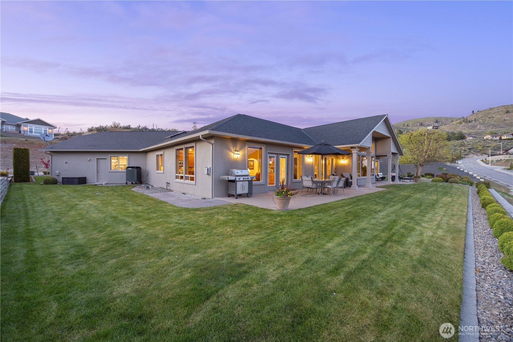 Stunning rear exterior showcasing expansive lawn space and a beautifully designed covered patio, all illuminated by warm evening lighting. Perfectly positioned to take in sweeping views of Lake Chelan and the surrounding mountains, this inviting outdoor setting is ideal for entertaining or relaxing at sunset.