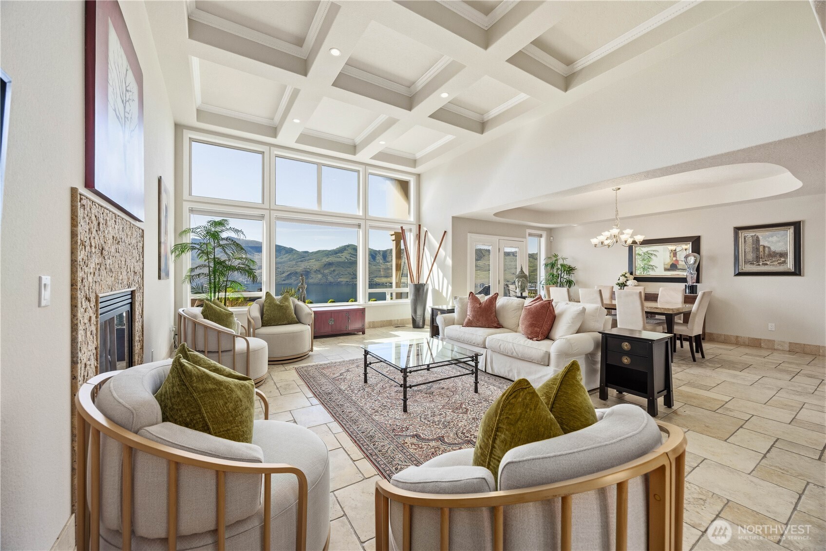 Stunning great room showcasing soaring coffered ceilings, expansive windows, and breathtaking panoramic views of Lake Chelan and the surrounding mountains. This light-filled living space features elegant tile flooring, a cozy fireplace, and an open flow into the dining area—perfect for both relaxed living and upscale entertaining, all framed by unforgettable scenery.